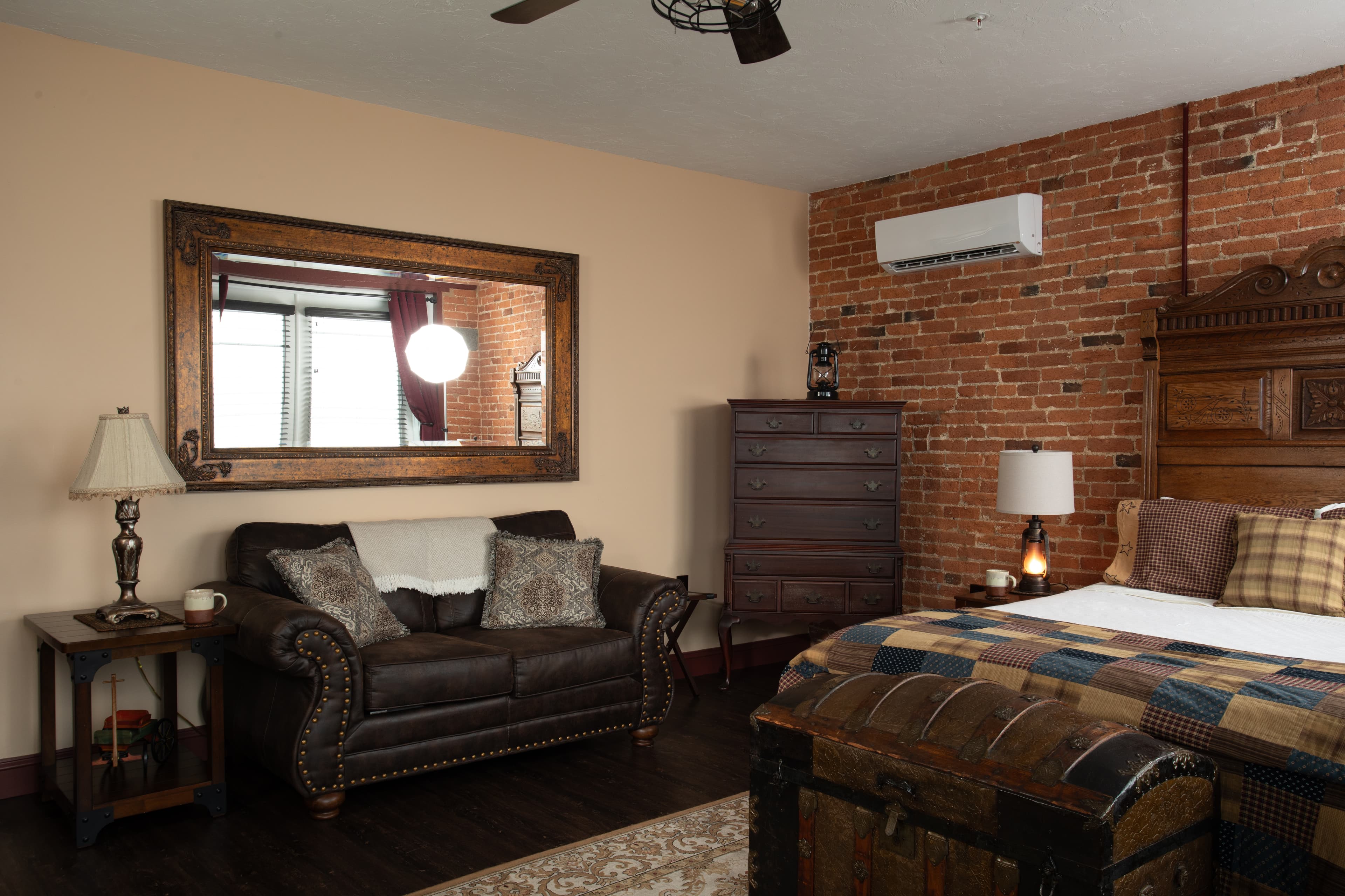 The Carriage House bedroom featuring a leather couch, wooden dresser, and a large mirror against a brick wall.