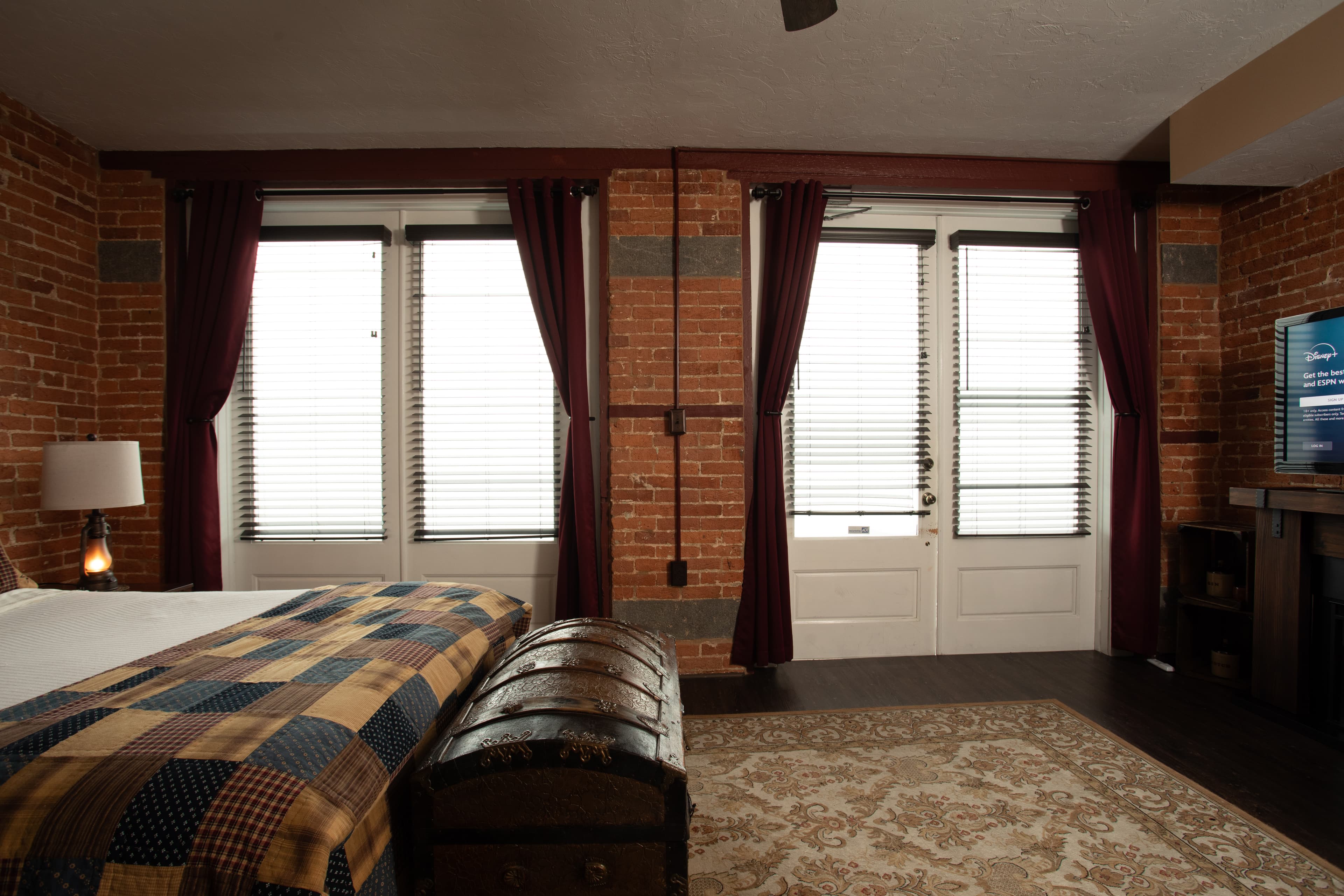The Carriage House bedroom featuring brick walls, dark curtains, a patterned blanket, and large windows.