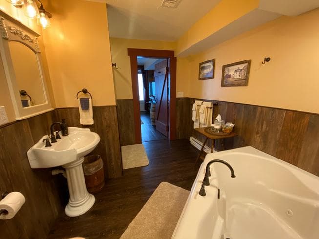 a bathroom with a white pedestal sink, a jetted bathtub, wood paneling on the lower walls, yellow paint on the upper walls, and a doorway leading to another room.
