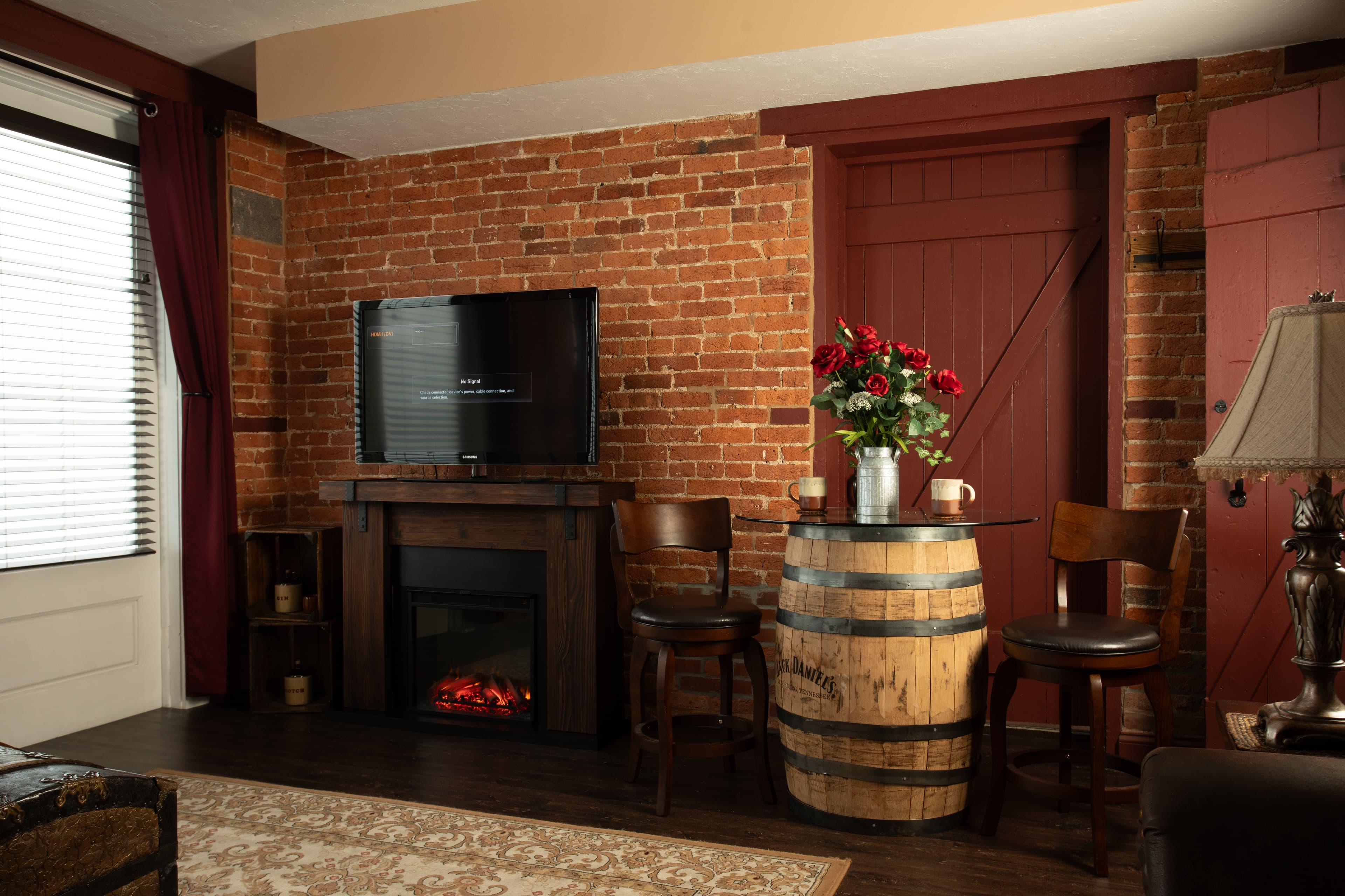 The Carriage House living room with a brick wall, a flat-screen TV, an electric fireplace, and a barrel table with flowers.