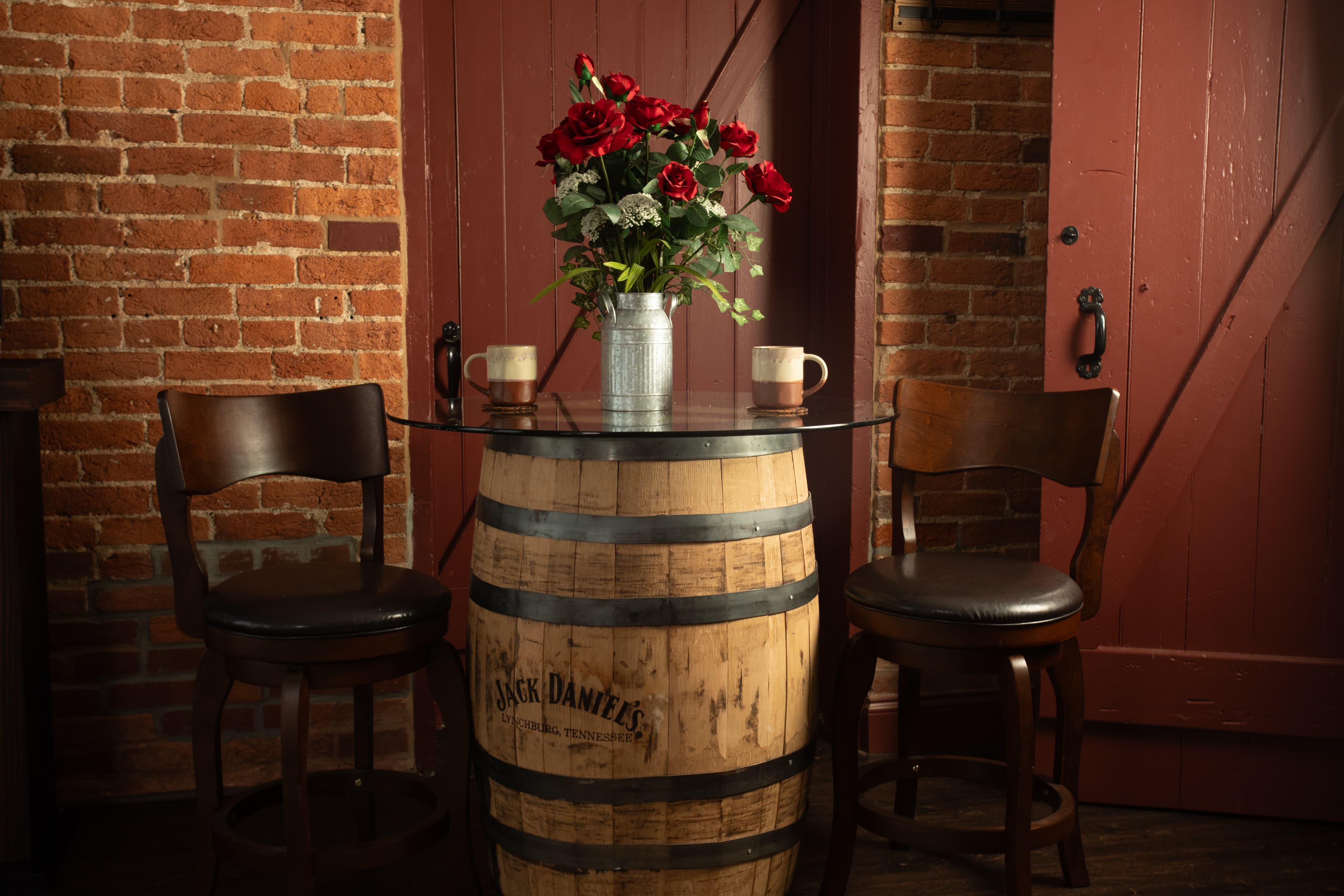 A rustic table made from a whiskey barrel topped with a glass surface, adorned with a vase of red roses and two mugs, set against a brick wall in the Carriage House.