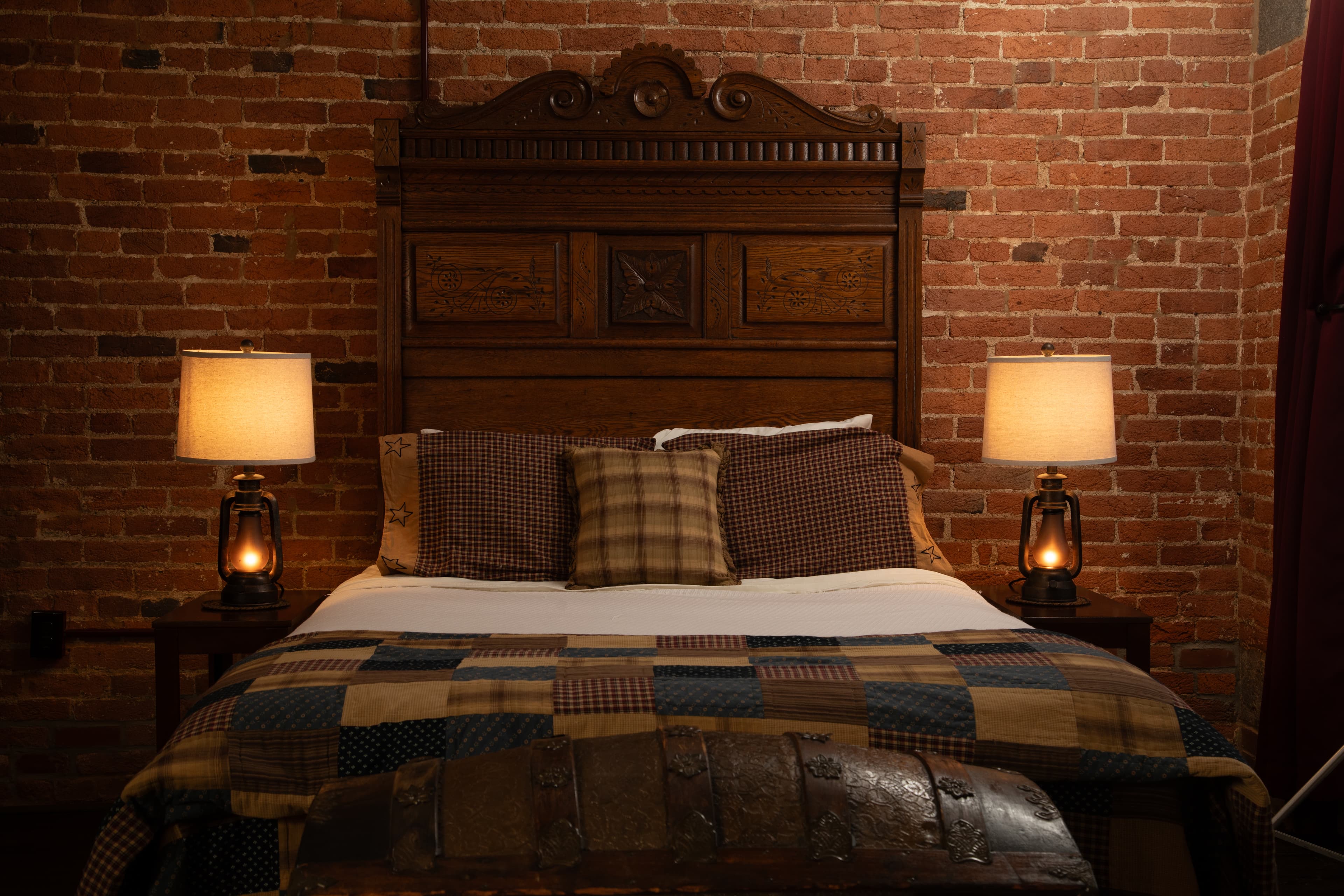 The Carriage House bedroom featuring a wooden bed, plaid bedding, and two bedside lamps against a brick wall.