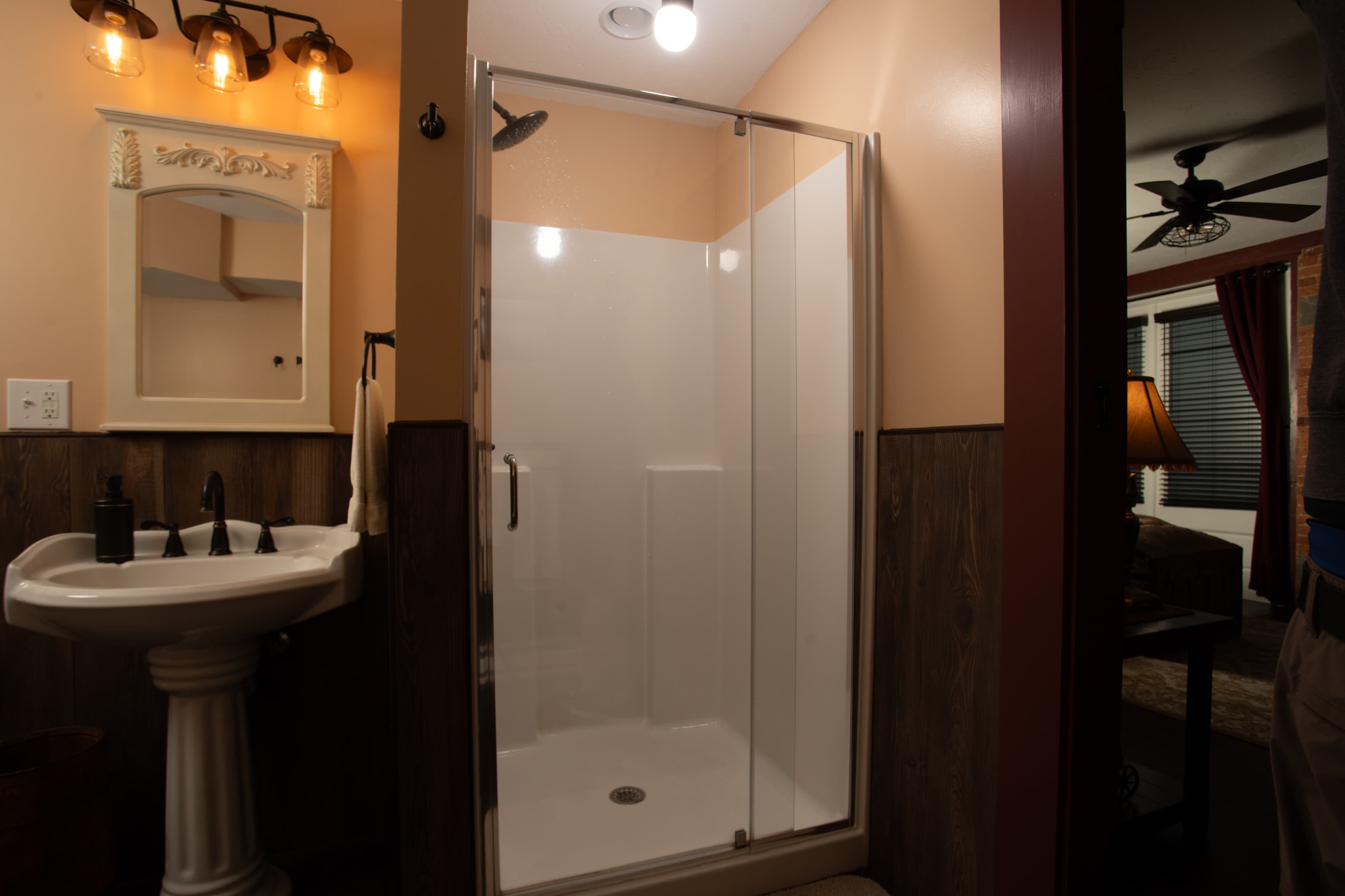 A modern bathroom in the Carriage House featuring a glass-enclosed shower, pedestal sink, and warm lighting.