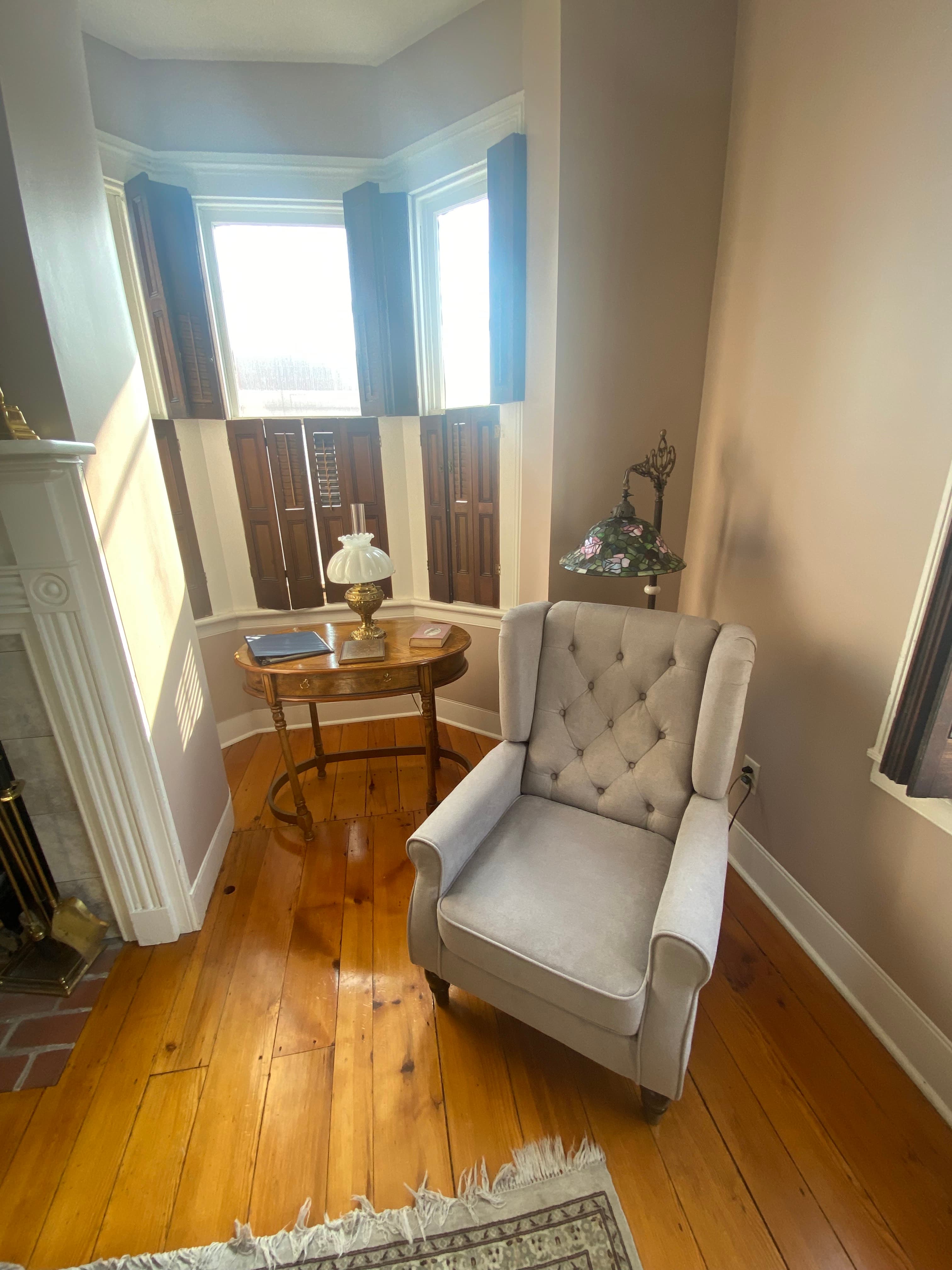 A light-colored armchair next to a bay window with striped curtains, a small table with a lamp, and wooden floors.