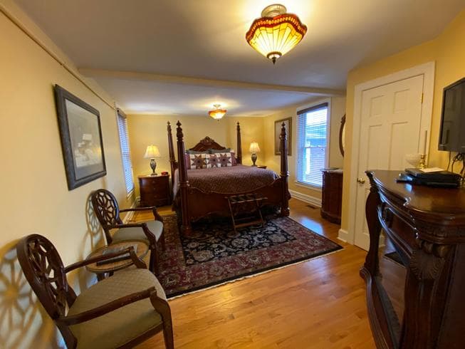 The Colonel Chamberlain Suite features a large ornate king-size wooden poster bed, 2 night stands with lamps, 2 chairs, and large wooden electric fireplace.
