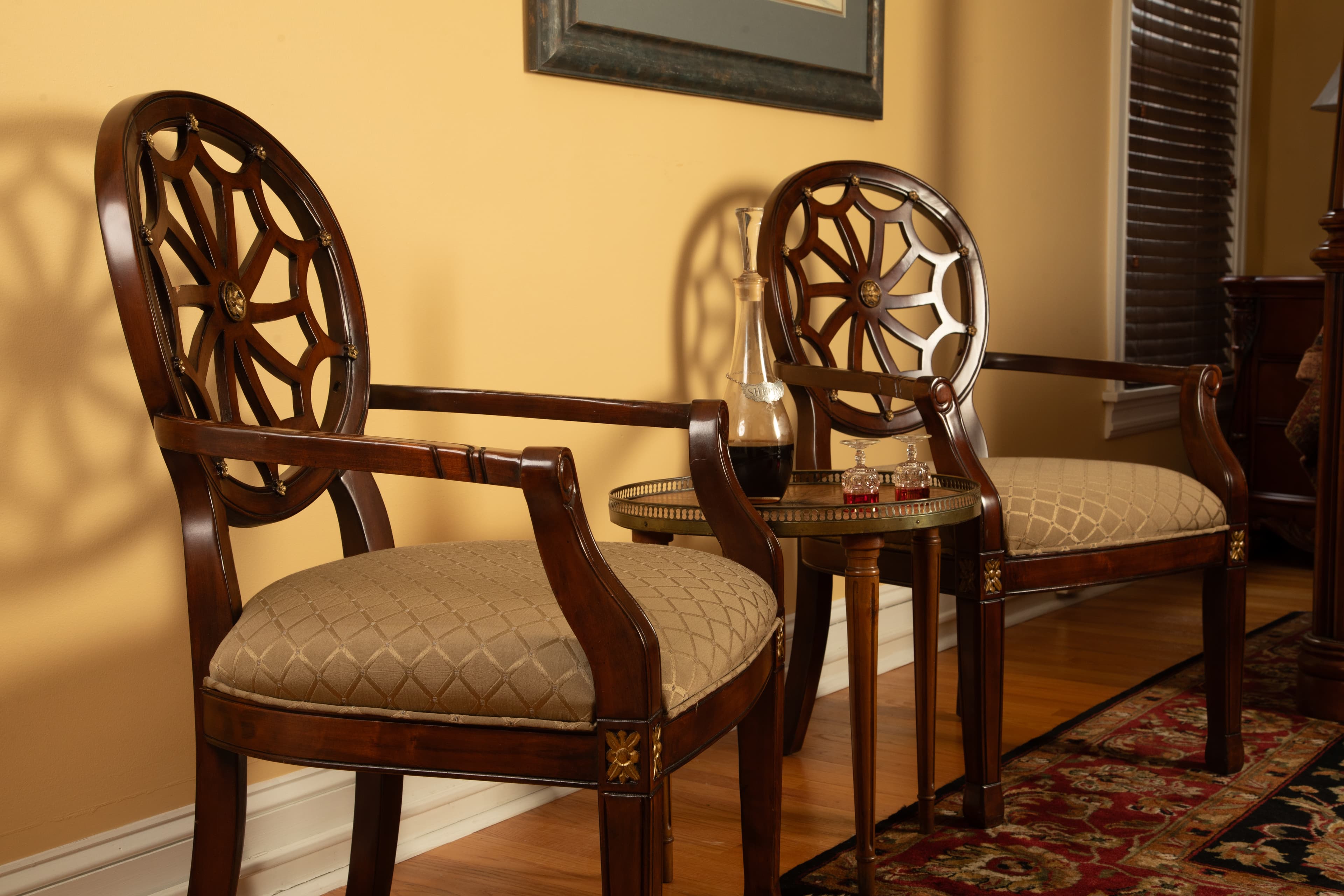 Two elegant wooden chairs with intricate back designs and cushioned seats, accompanied by a small round table and decorative items against a warm wall in the Chamberlain bedroom.