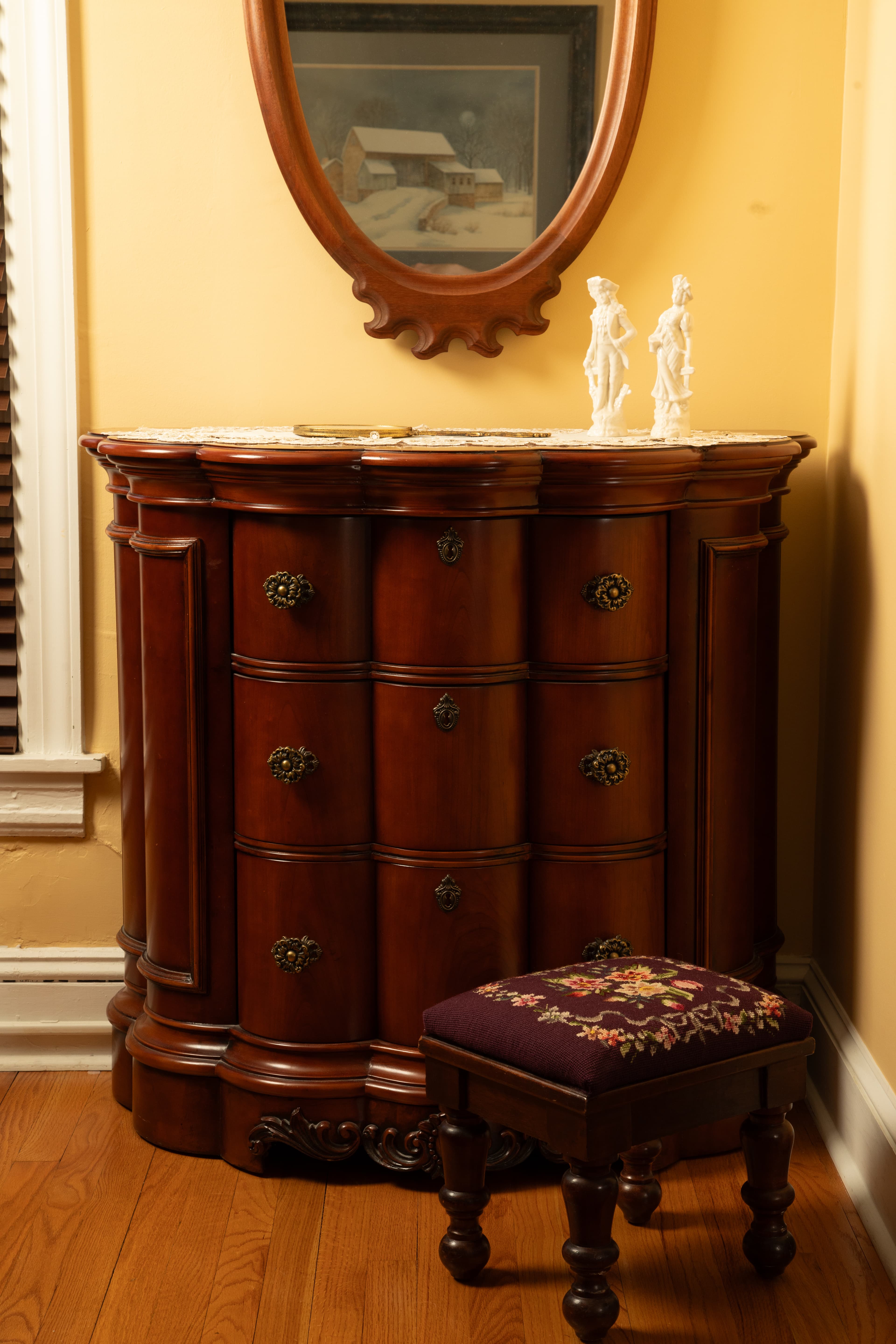 A polished wooden chest of drawers with intricate hardware and a small upholstered stool beside it, set against a warm yellow wall in the Chamberlain bedroom.