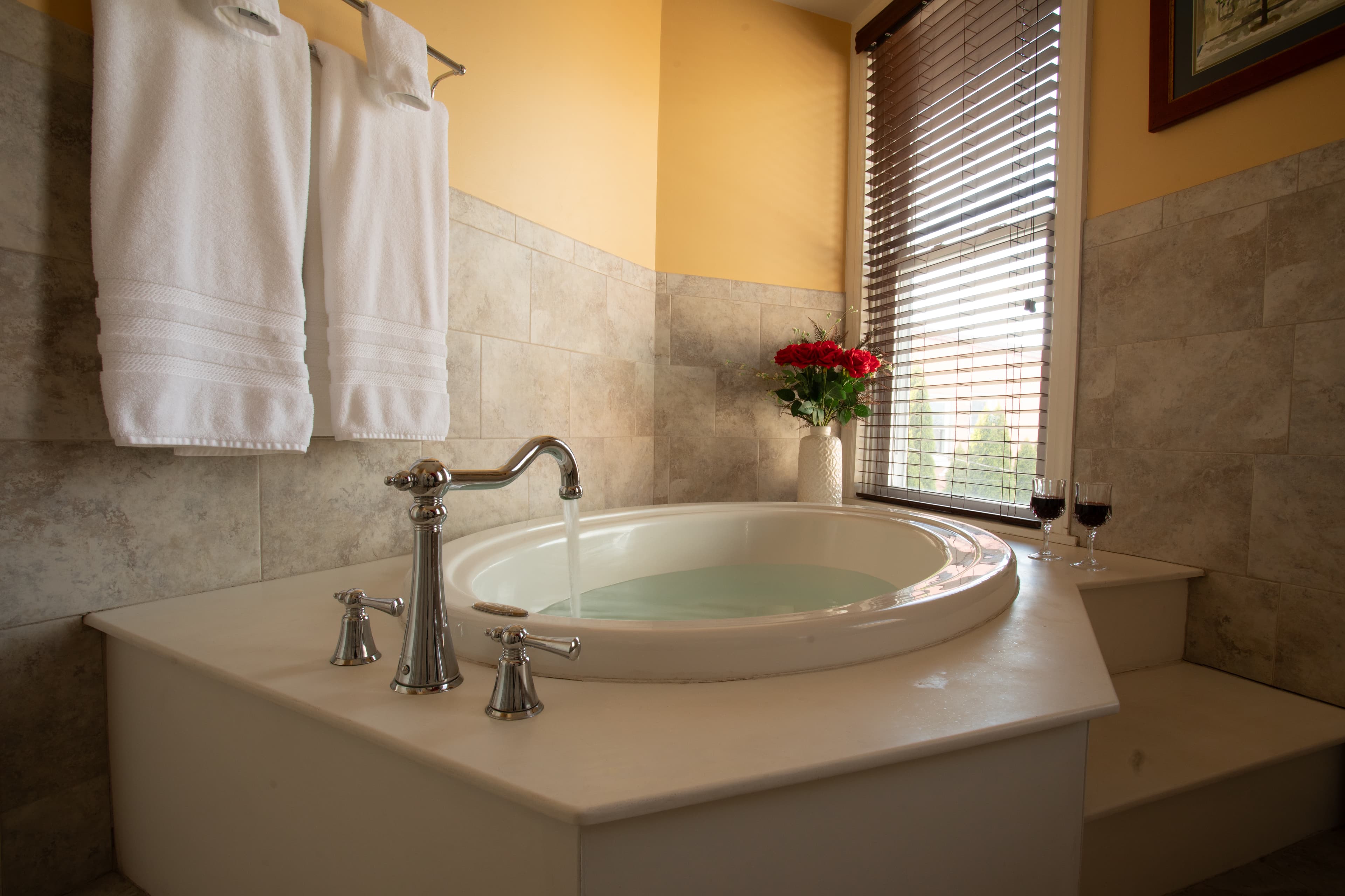 A luxurious bathroom in the Chamberlain featuring a round soaking tub, fresh roses, and two wine glasses by a window.