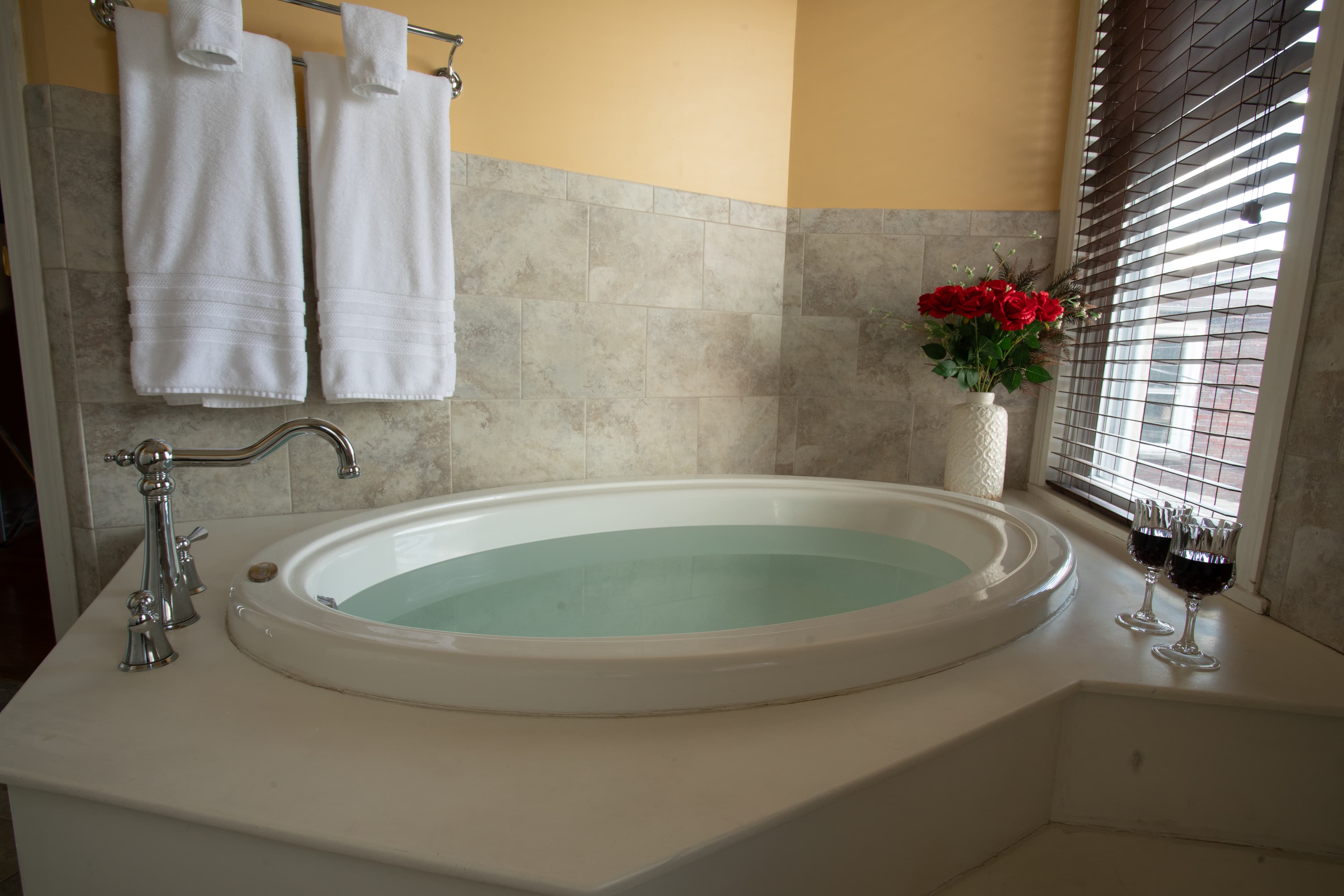 A stylish bathroom in the Chamberlain featuring an oval soaking tub, white towels, flowers, and two glasses of wine.