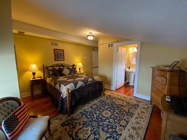The Lt. Pohlman Chamber features a queen-size metal bed, 2 night stands with lamps, a dresser, 2 chairs, an electric stove, and original wooden floors with area rug.