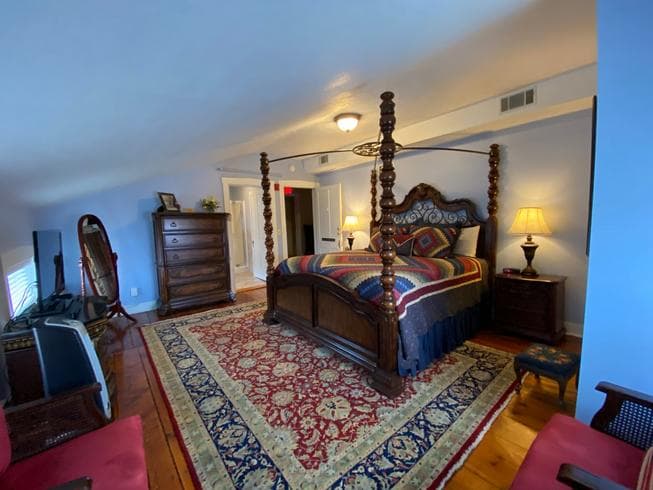 The Major General Meade Suite features a Queen Size Wooden Four Poster Bed, 2 night stands with lamps, a dresser, 2 chairs, an electric fireplace, TV, and original wooden floors with area rug.