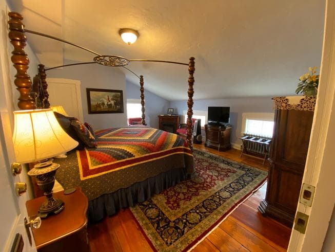 The Major General Meade Suite features a Queen Size Wooden Poster Bed, 2 night stands with lamps, a dresser, 2 chairs, an electric fireplace, TV, and original wooden floors with area rug.