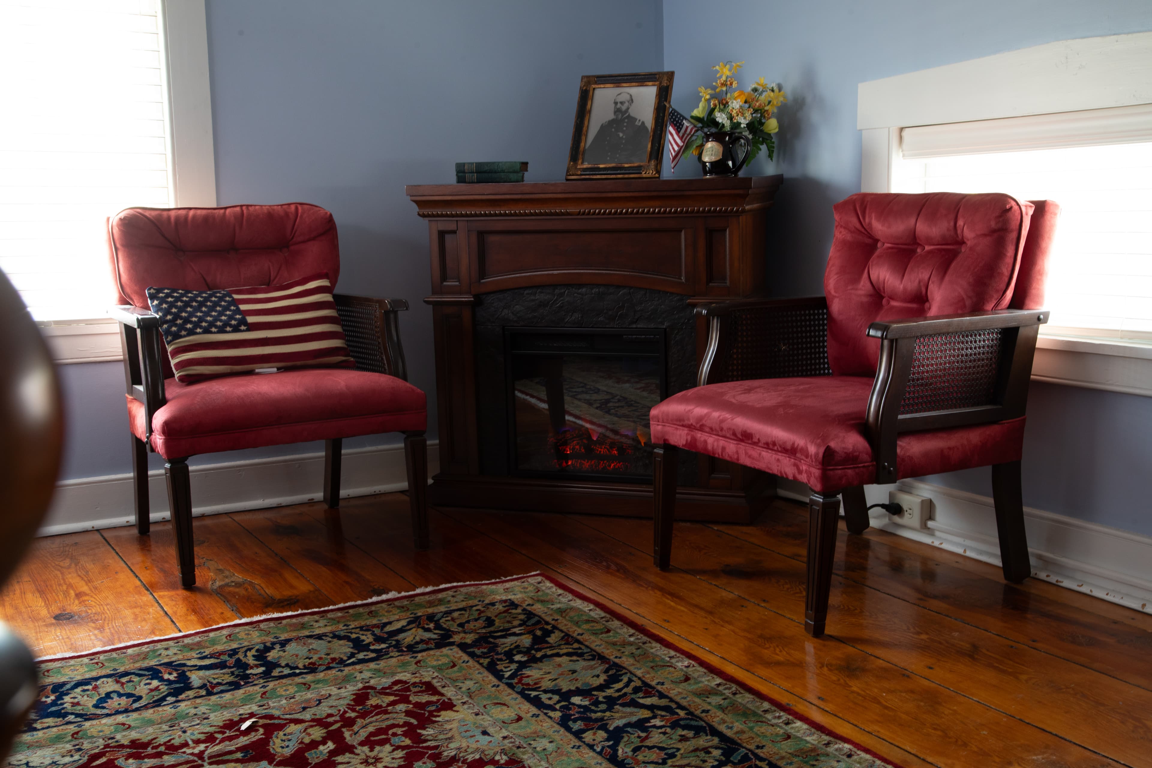 A cozy room featuring two red chairs, a decorative fireplace, and a small bookshelf with a portrait and flowers.
