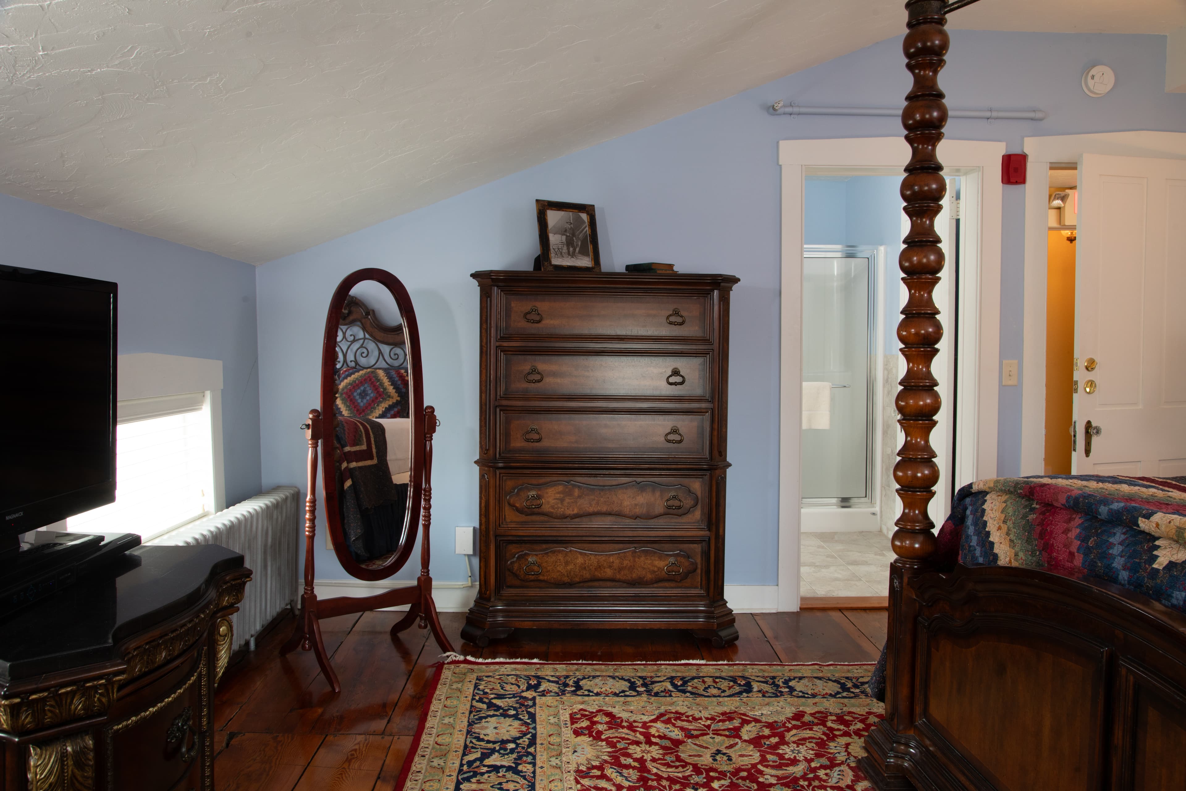 A cozy attic bedroom featuring a wooden dresser, a full-length mirror, and a doorway to a bathroom.