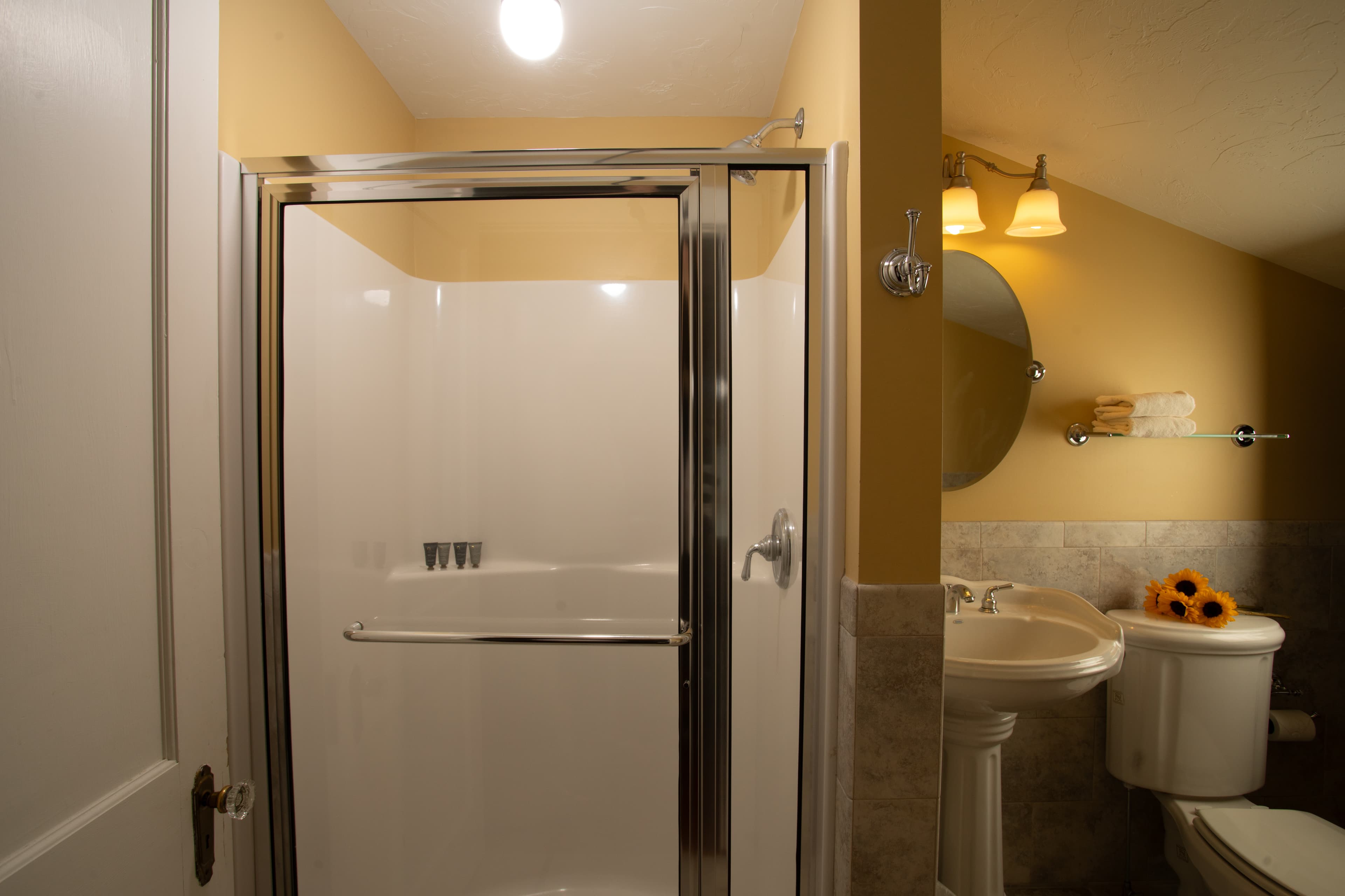 A small bathroom featuring a glass shower, a sink with a mirror, and a toilet against yellow walls.
