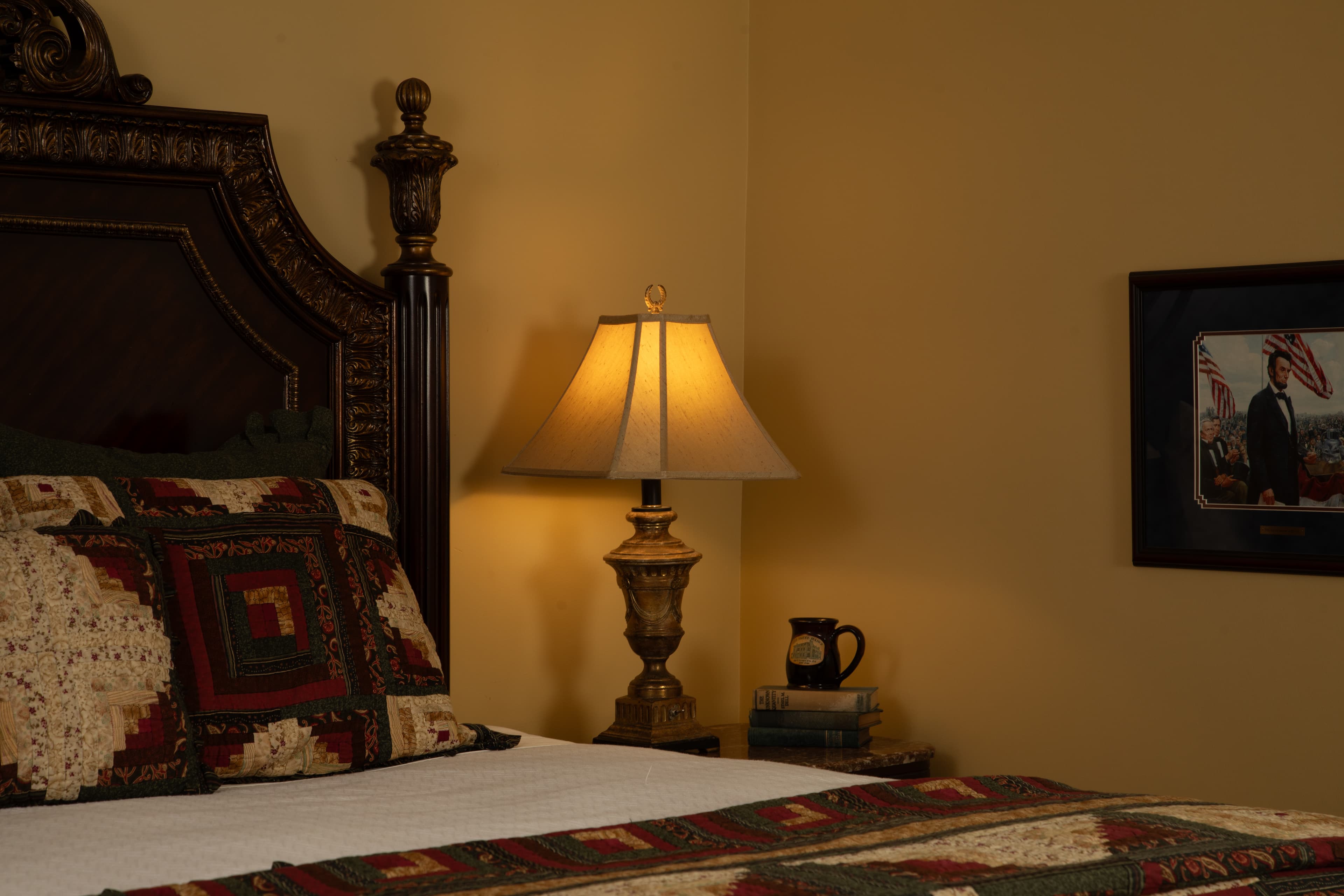 A cozy bedroom featuring a patterned quilt, a bedside lamp, and a framed picture on the wall.