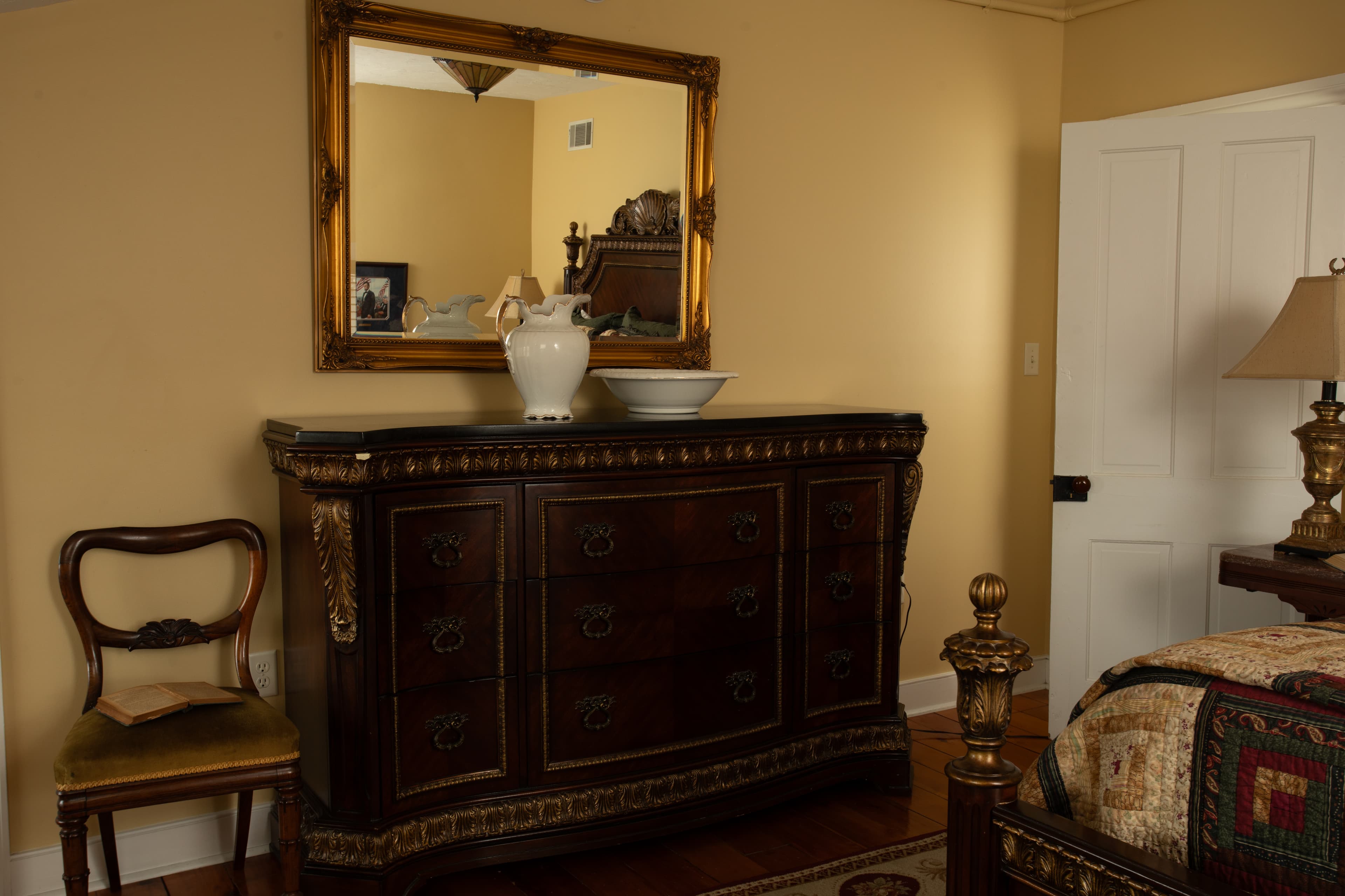 A wooden dresser with decorative details, a mirror above, and a vase sits in a warmly colored bedroom.