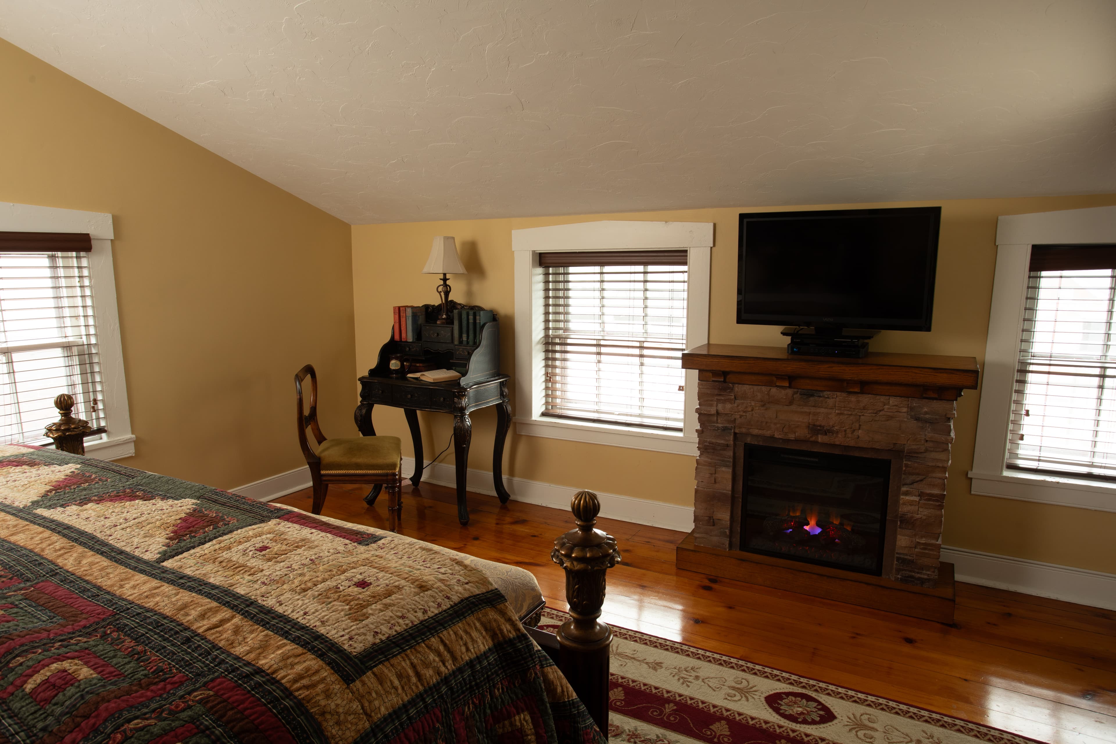 Cozy bedroom featuring a quilted bed, a fireplace, a TV, and a writing desk.