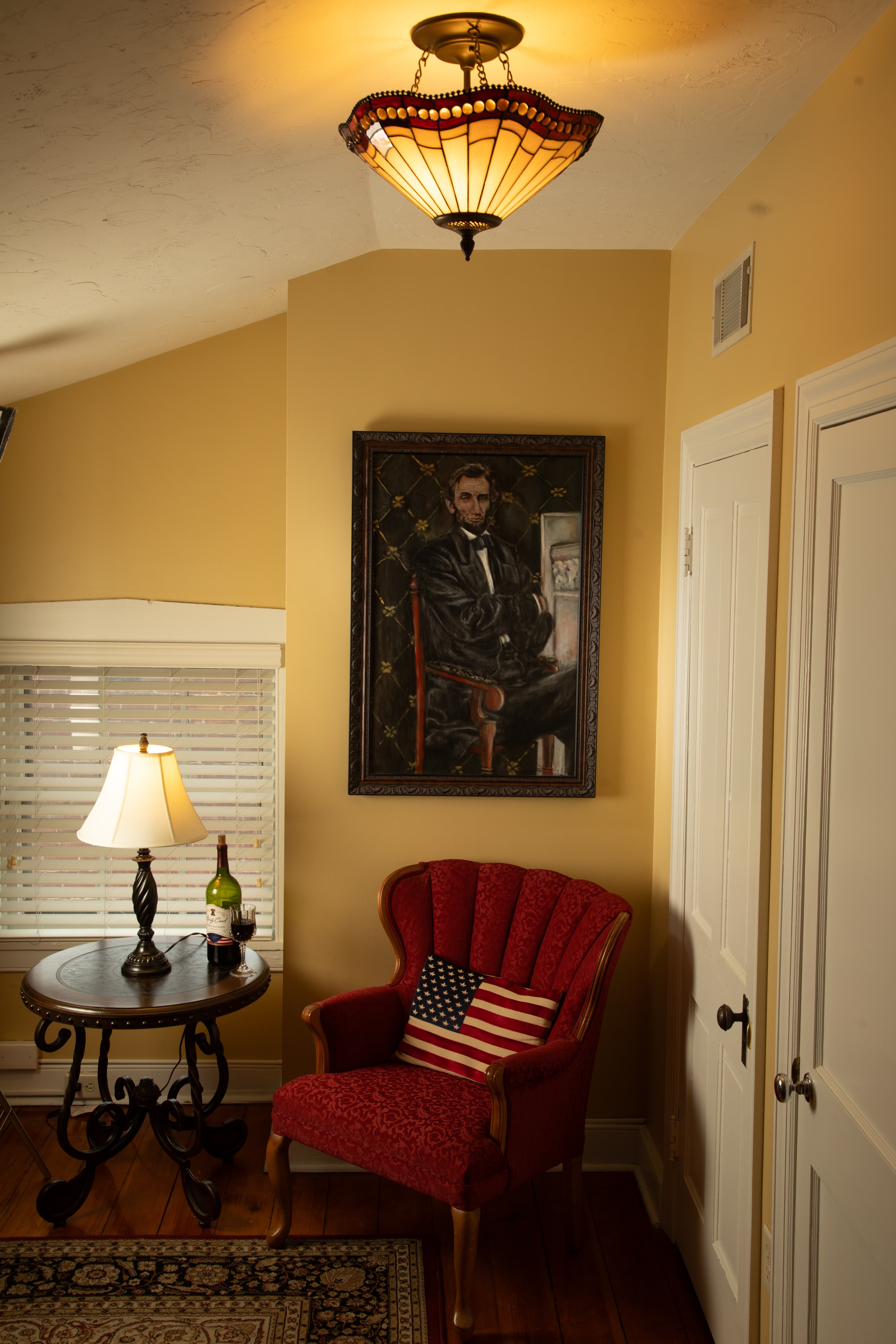 A cozy corner featuring a red upholstered chair, a side table with a lamp and wine, and a portrait of Abraham Lincoln on the wall.