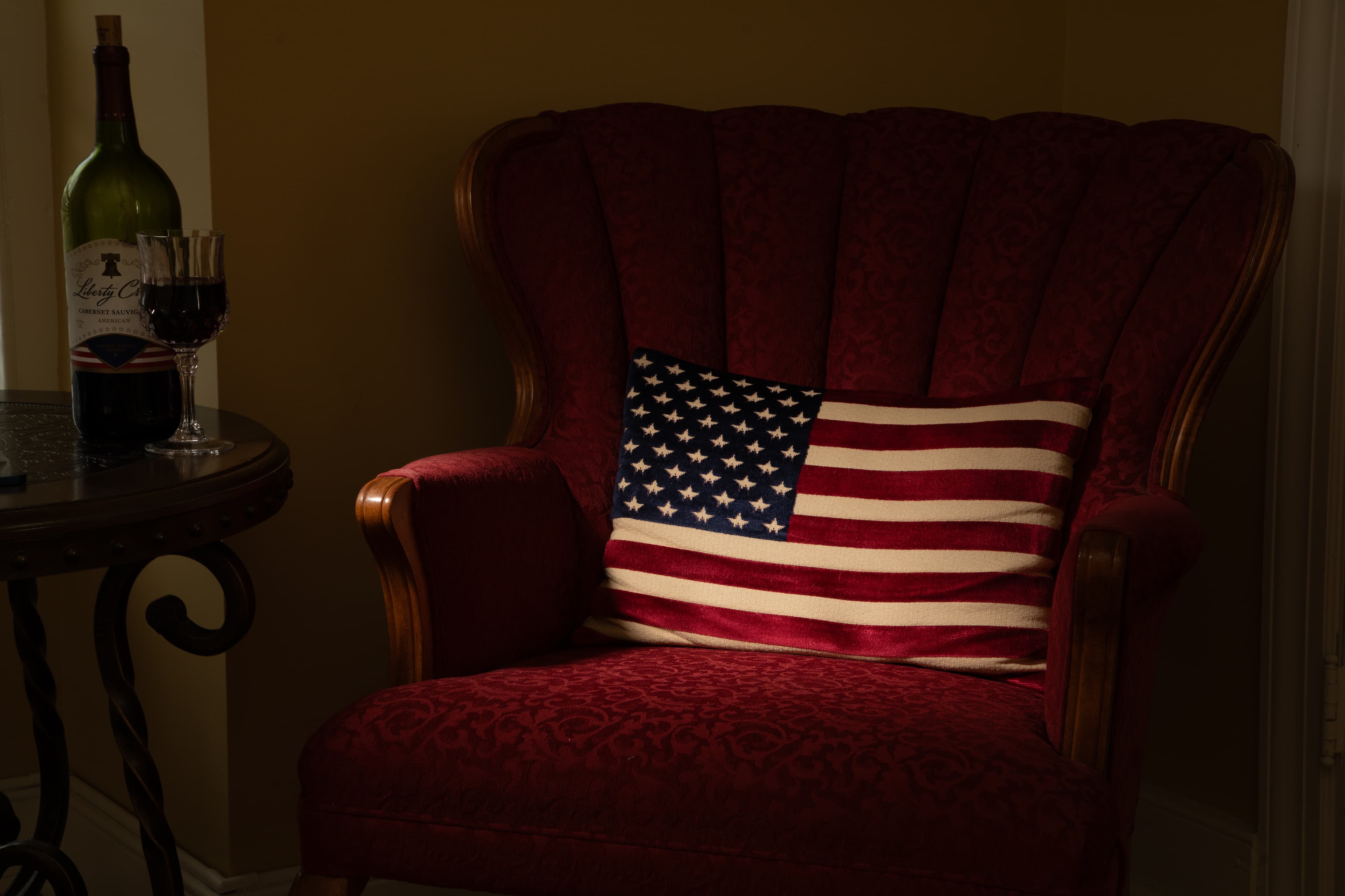 A red armchair with a striped American flag pillow next to a table holding a wine bottle and glass.