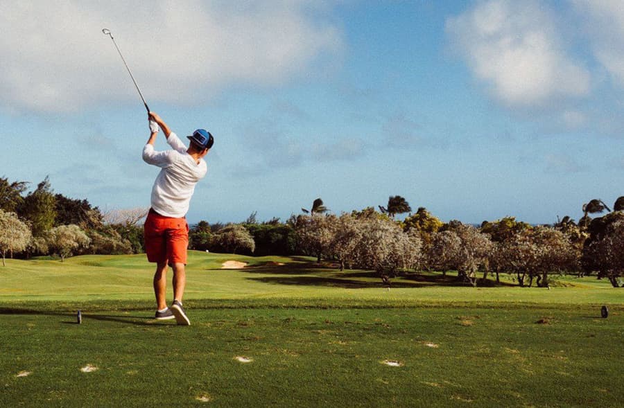 A golfer swings a club on a lush green course under a blue sky.