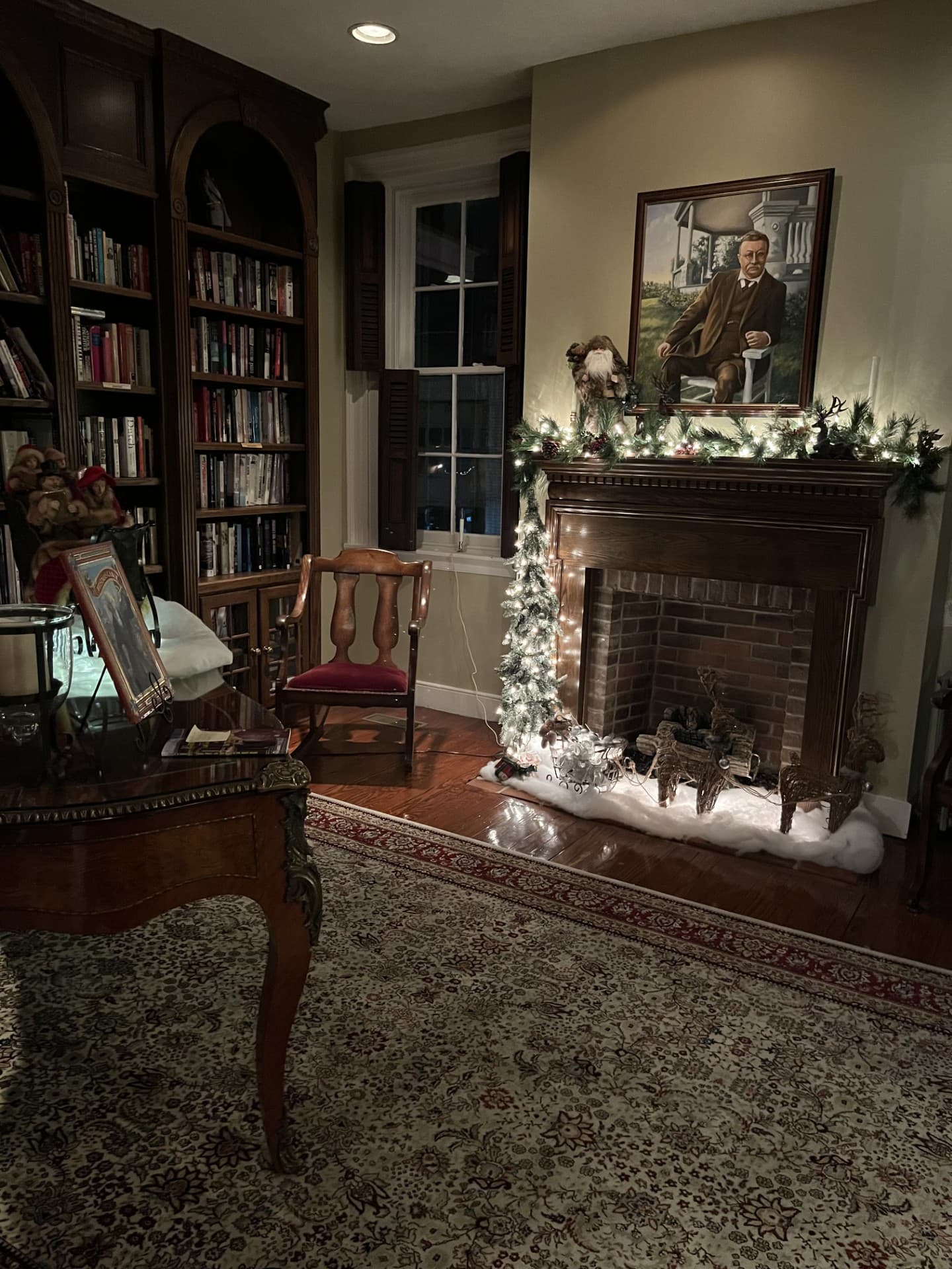 Cozy room featuring a bookshelf, armchair, and a festive fireplace with a small tree and holiday decorations.
