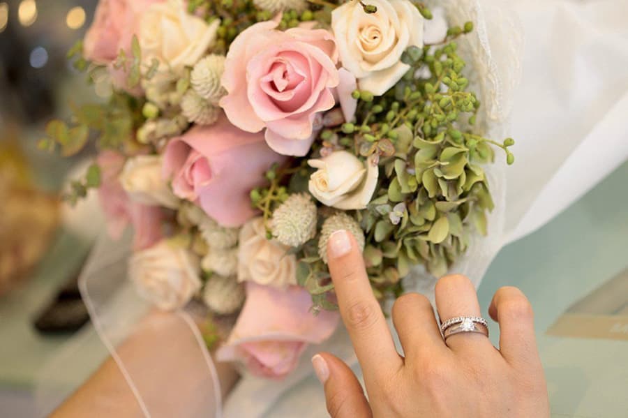A hand with a silver ring points toward a bouquet of light pink roses and greenery.