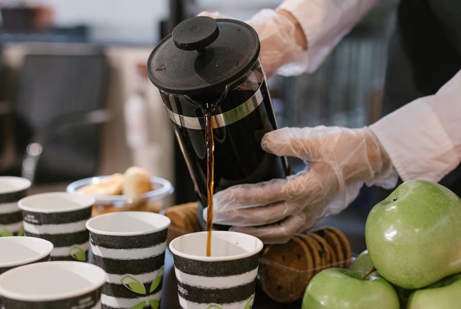 A person in gloves pours coffee from a French press into striped cups, accompanied by green apples and cookies on a table.