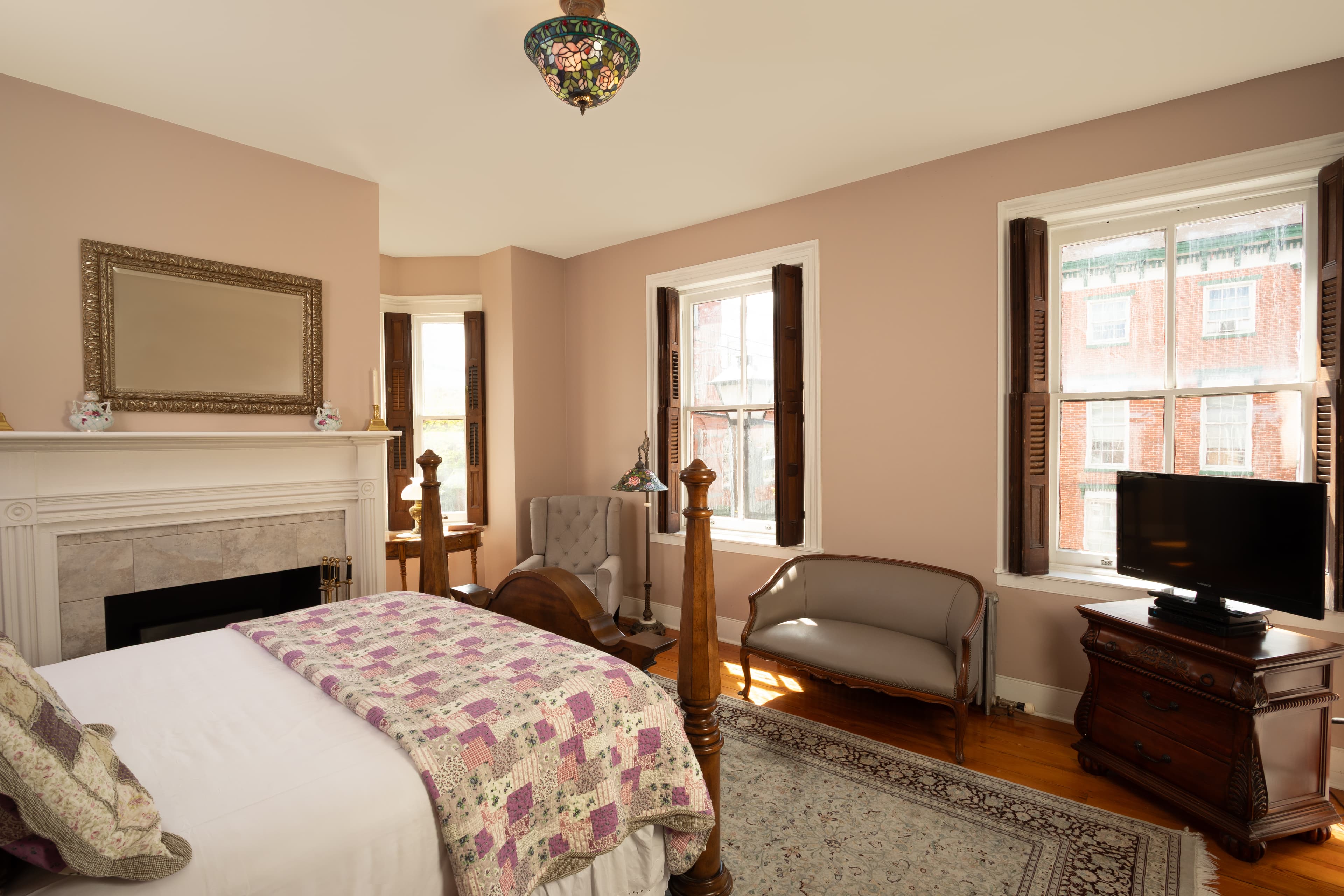 A well-decorated bedroom featuring a bed with a quilt, a vintage armchair, a media console, and large windows.