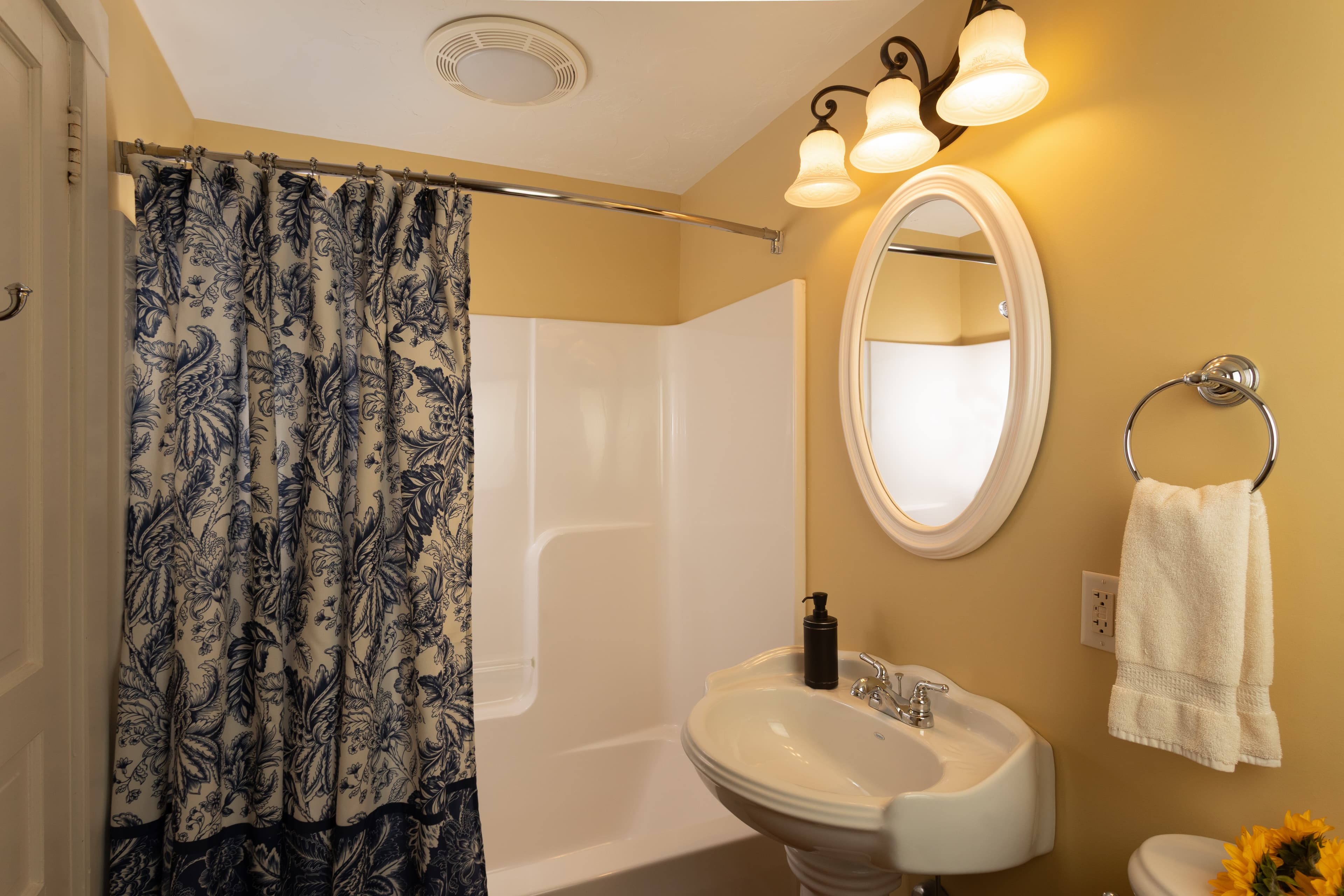 A small bathroom featuring a shower with a floral curtain, a sink, a round mirror, and decorative lighting.