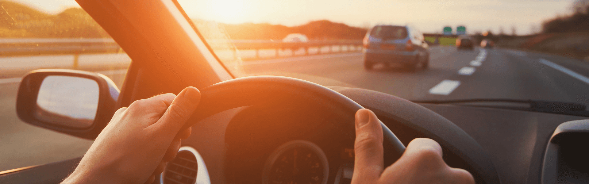 Hands gripping a steering wheel while driving on a sunlit highway.