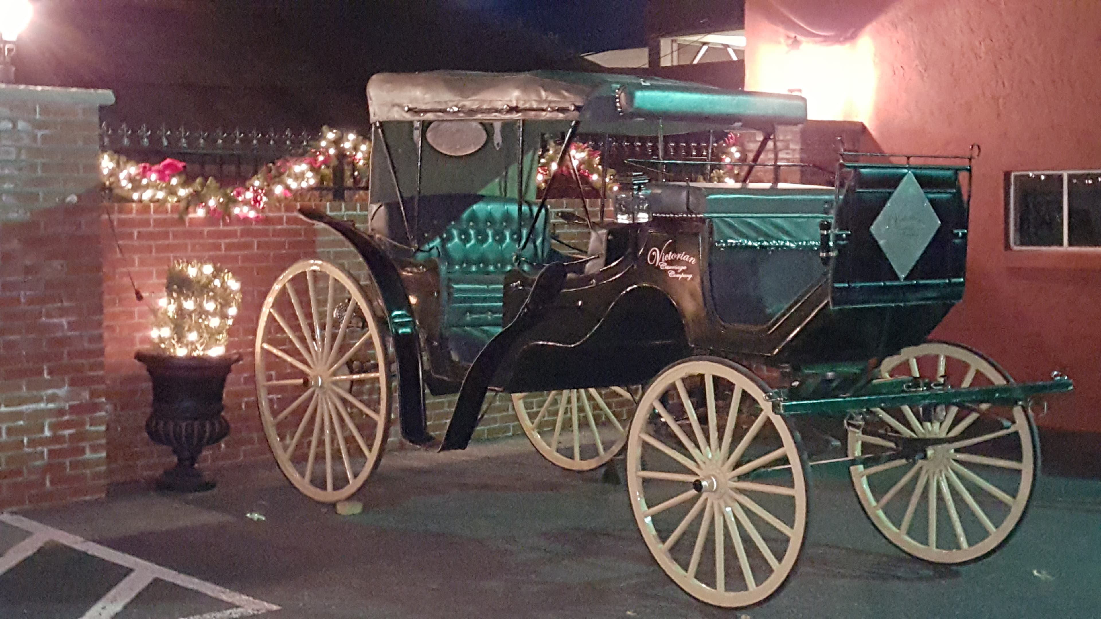 A vintage black carriage decorated with festive lights is positioned beside a brick wall.