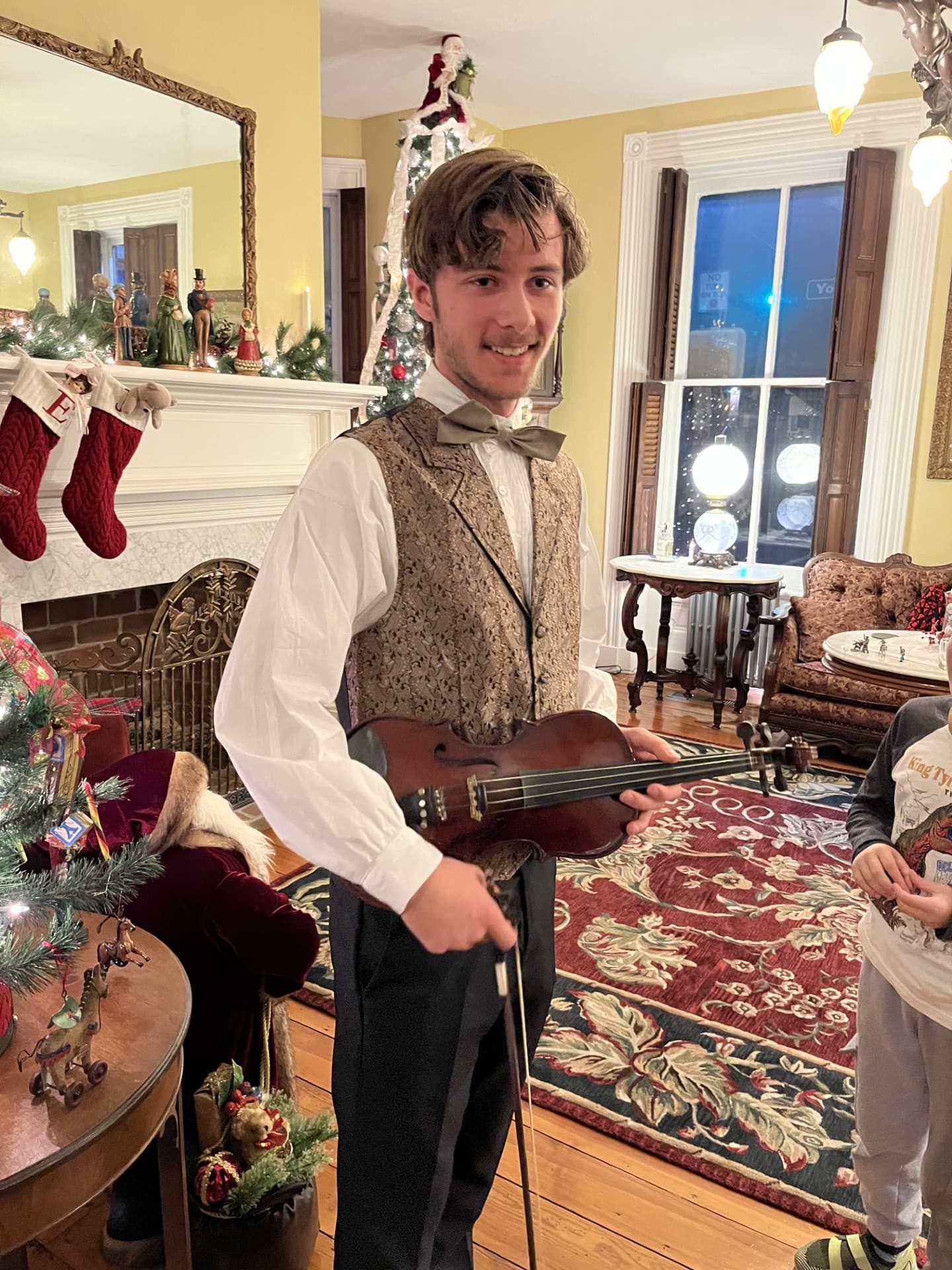 A young man in formal attire holds a violin in a festive room decorated for the holidays.
