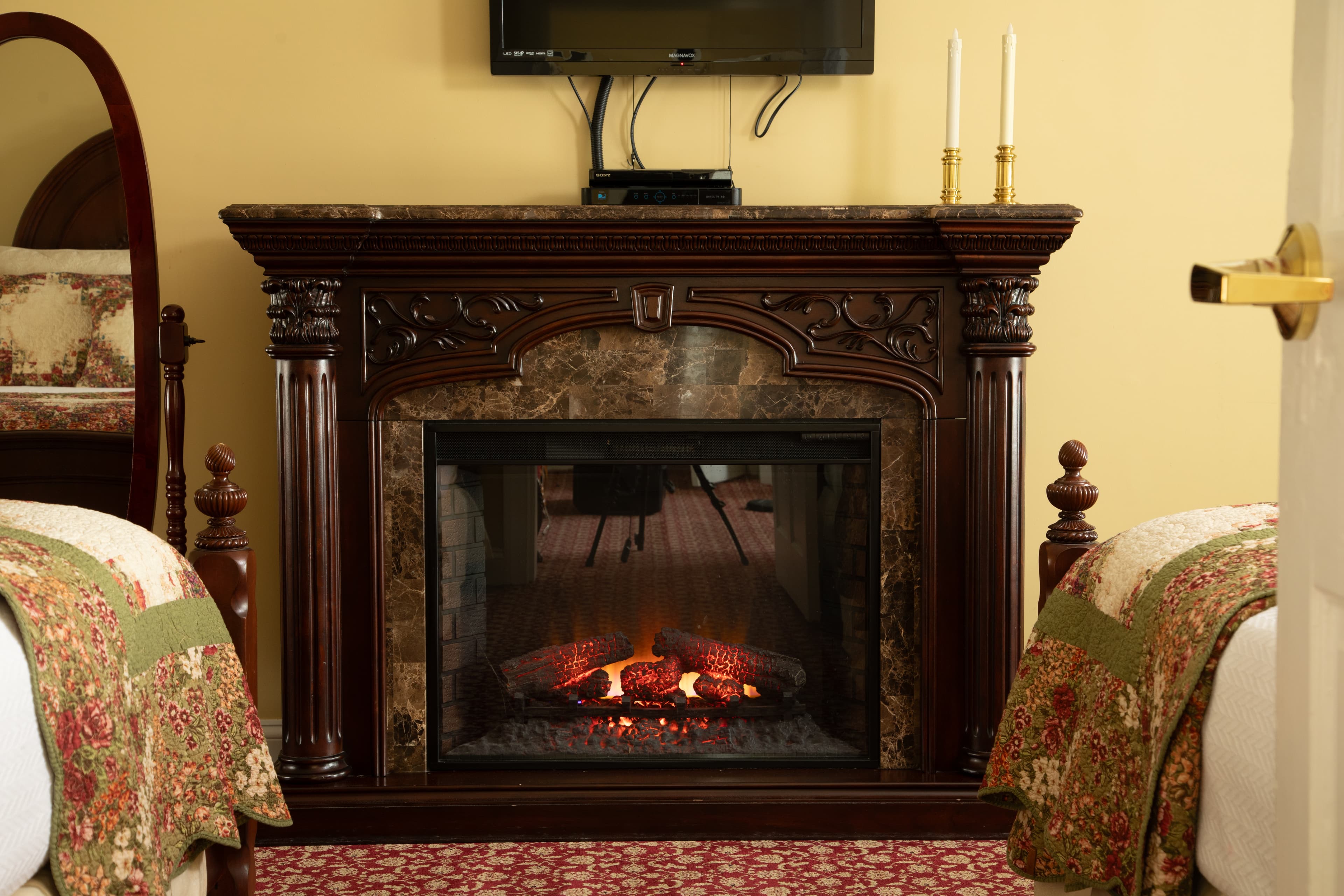 A sitting area with yellow walls, a red A cozy electric fireplace with a decorative mantle and a television above it, flanked by two beds.carpet, a Victorian sofa, a lamp, a framed picture, and a doorway leading to another room.