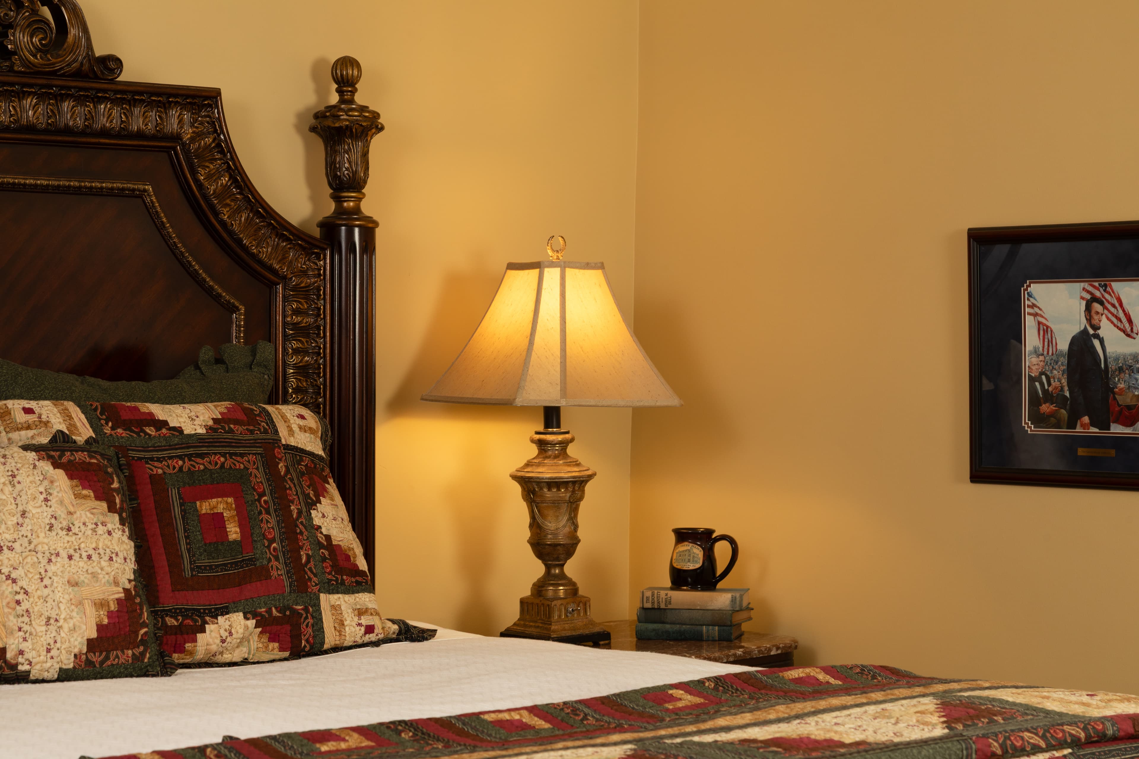 A cozy bedroom featuring a patterned quilt, a bedside lamp, and a framed picture on the wall.