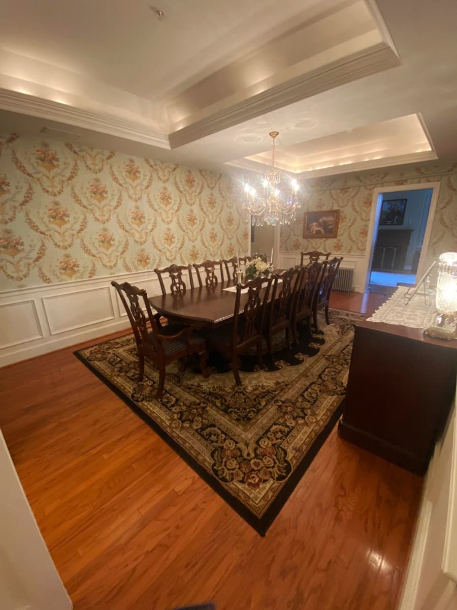 A spacious dining room featuring a long wooden table, elegant chairs, floral wallpaper, and a chandelier.