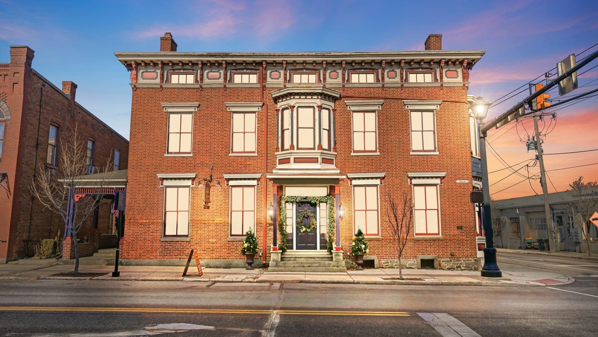 A historic red brick building with decorative trim, featuring floral decorations at its entrance and a sunset backdrop.