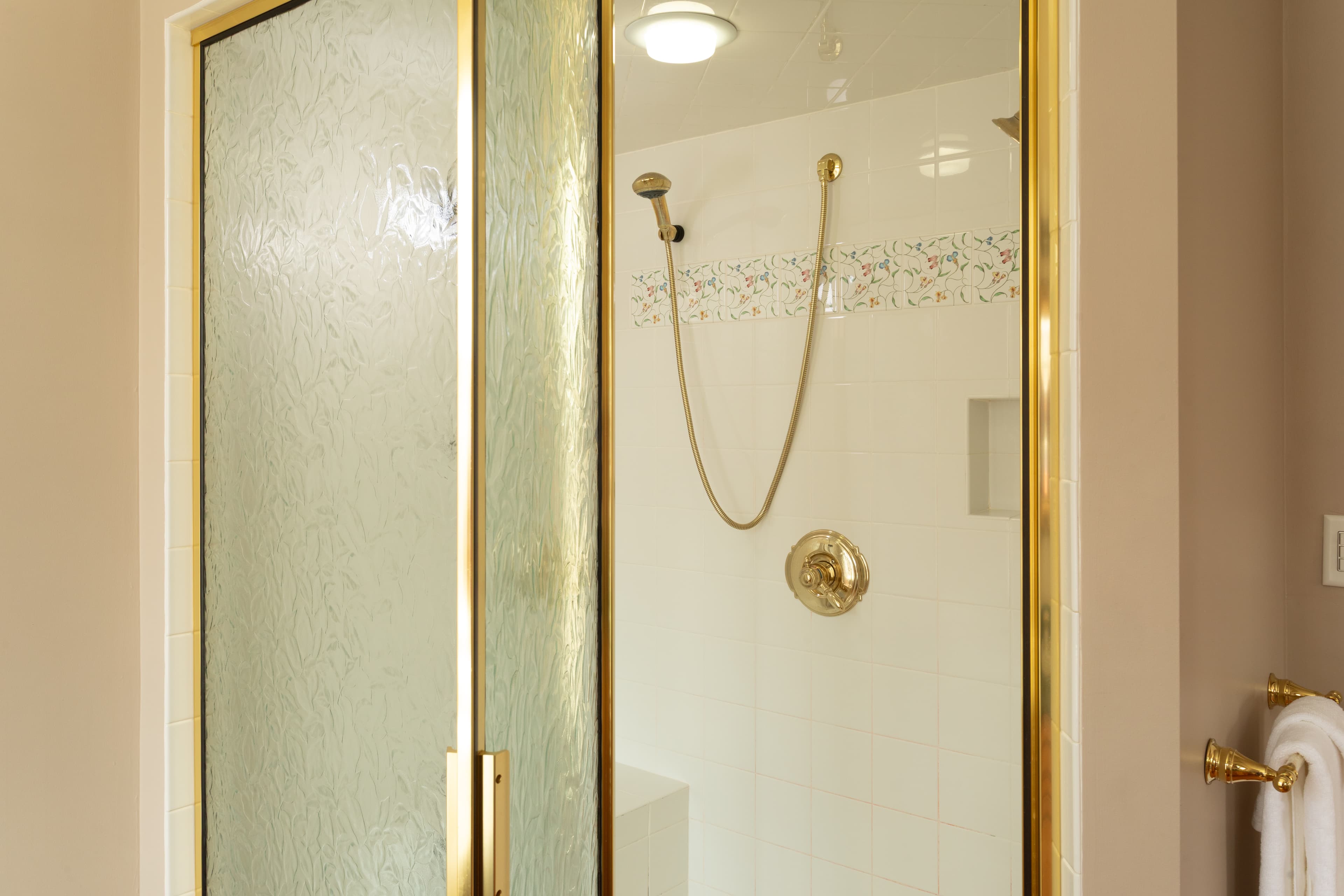 A glass shower stall with gold trim features a handheld showerhead and tiled walls.