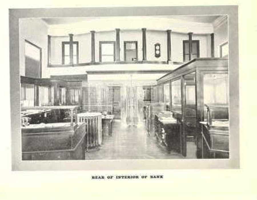 Historic black-and-white photograph of the interior of a bank, featuring wooden counters and large windows.