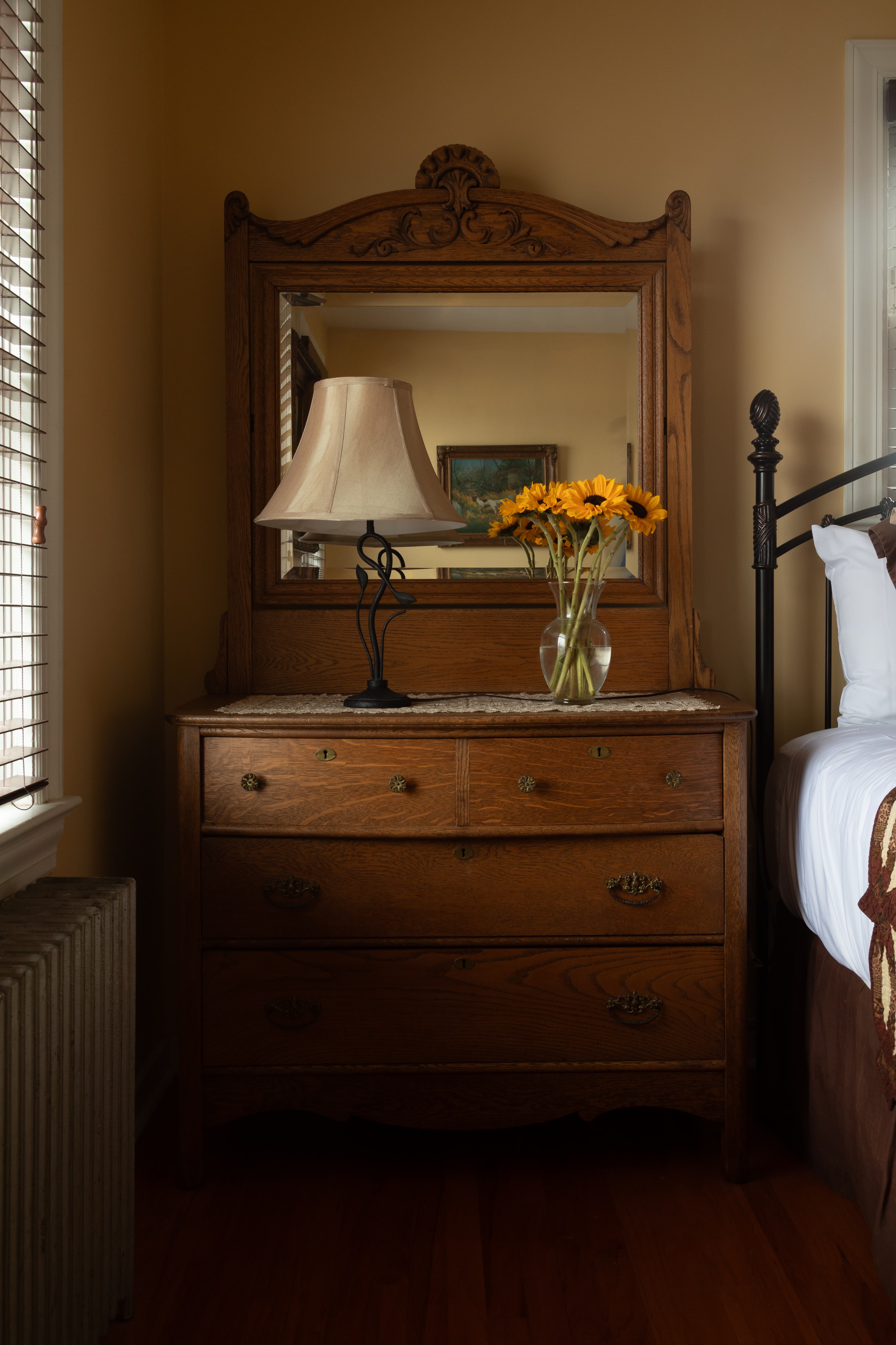A vintage wooden dresser with a mirror, a lamp, and a vase of yellow flowers.