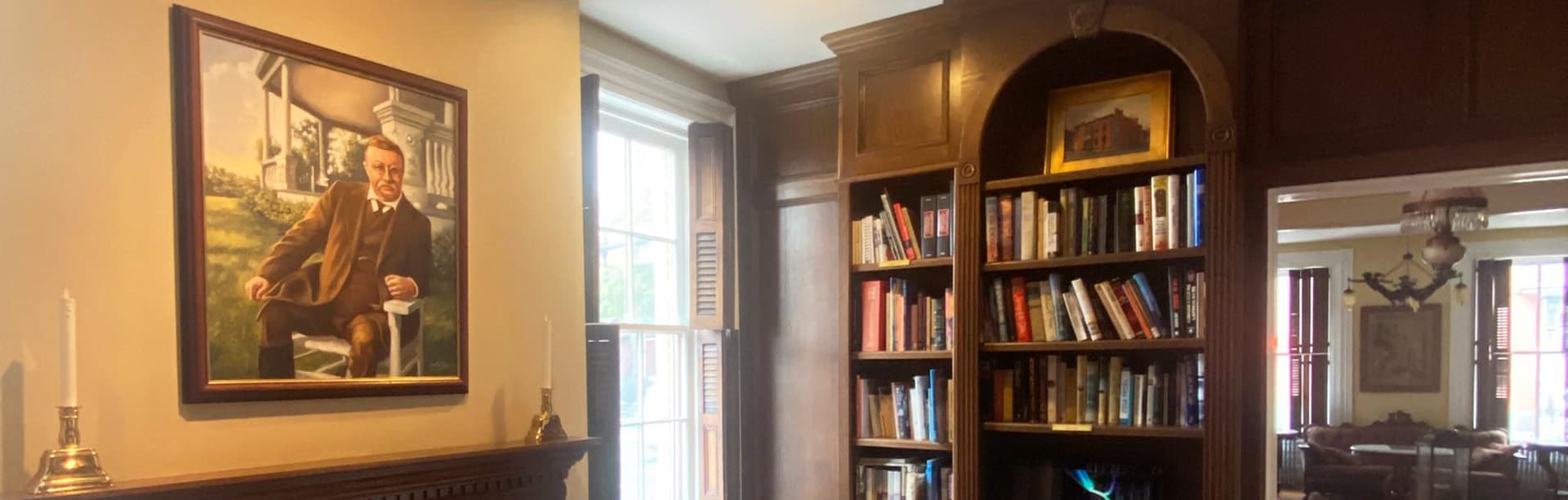 A room featuring a portrait of a man next to a bookshelf filled with books and a window.