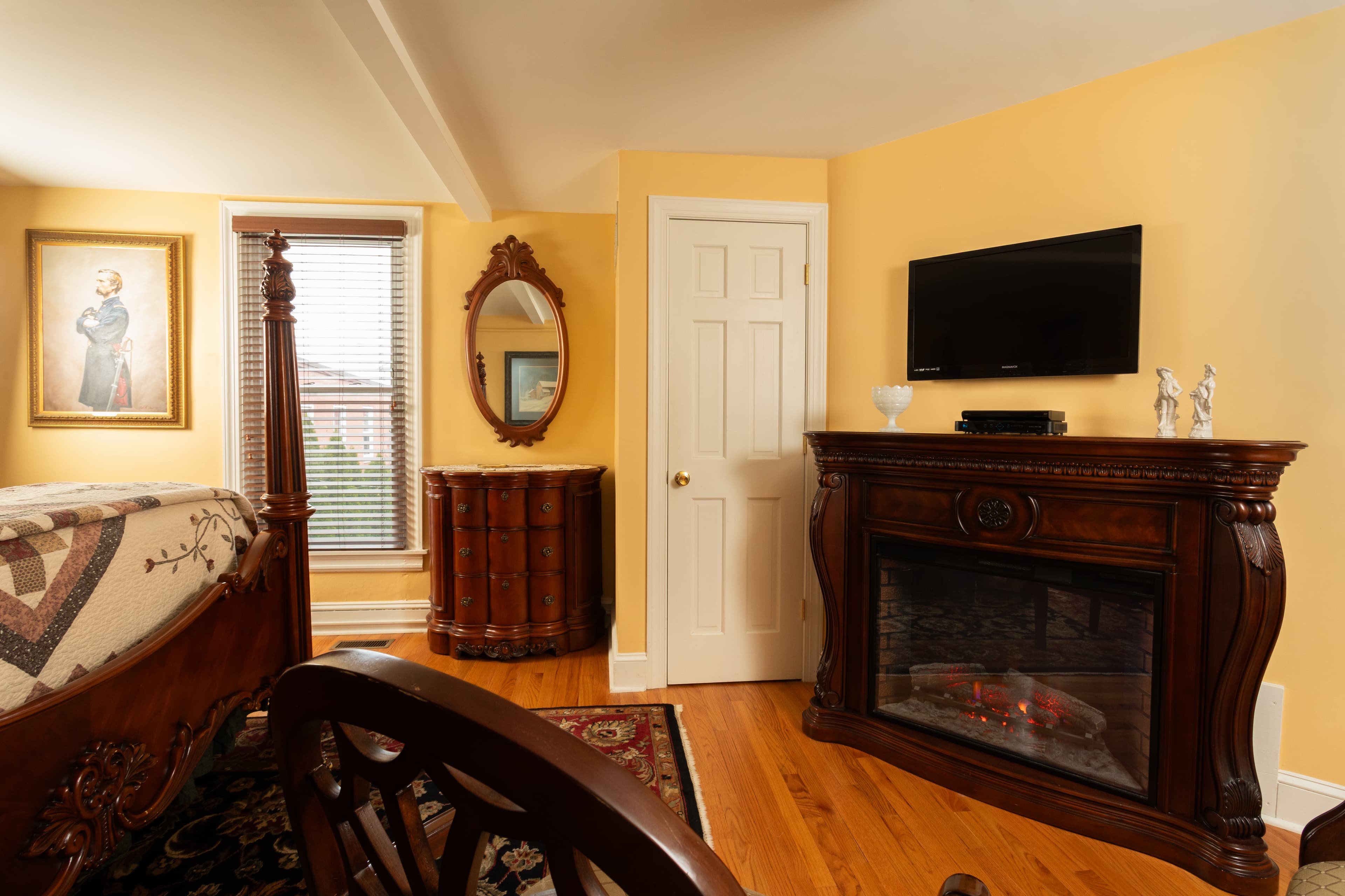 Cozy bedroom featuring a wooden bed, dresser, fireplace, and a wall-mounted TV against a warm, yellow backdrop.