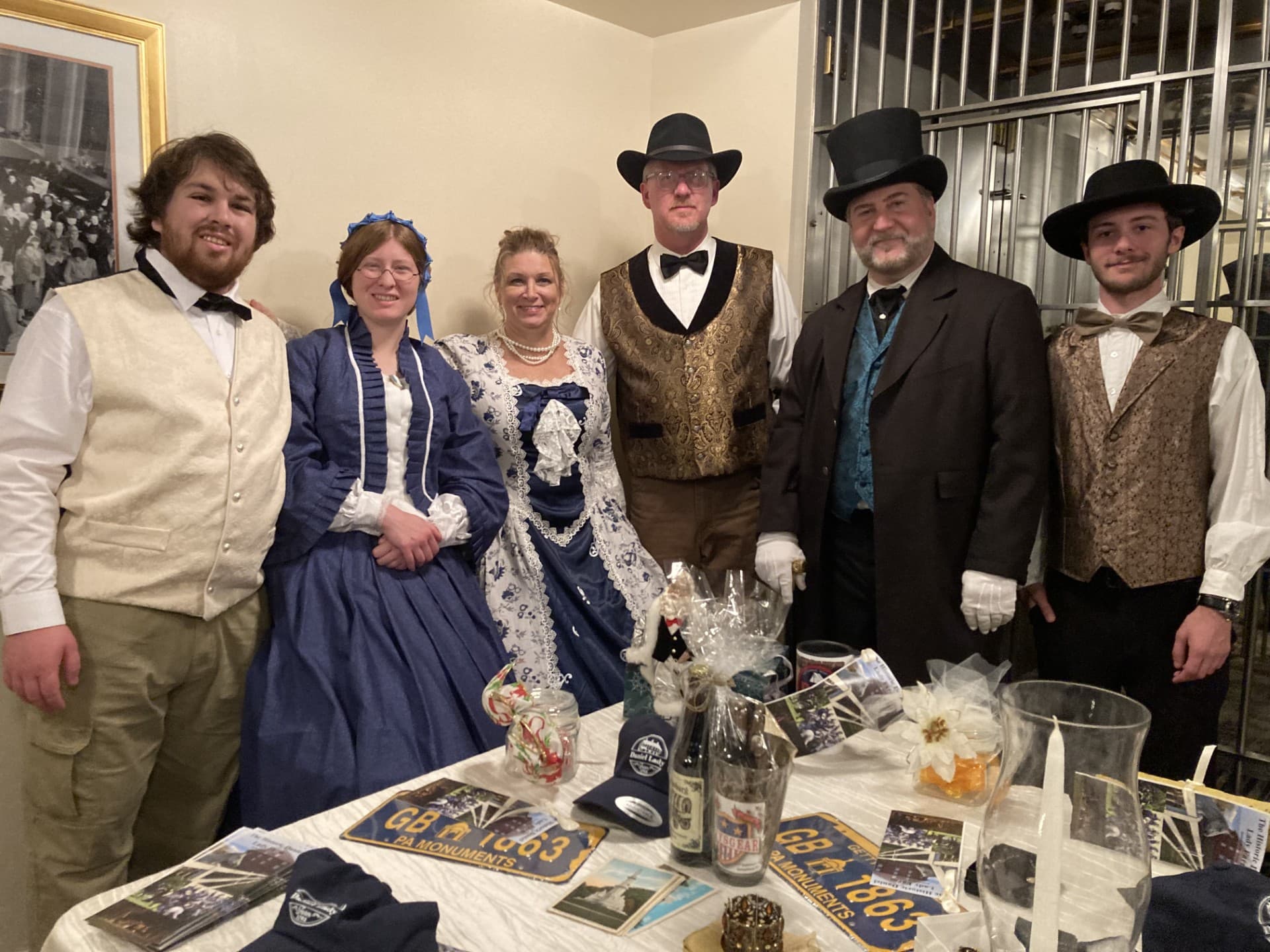A group of six people in vintage formal attire poses together at a table adorned with decorations and memorabilia.