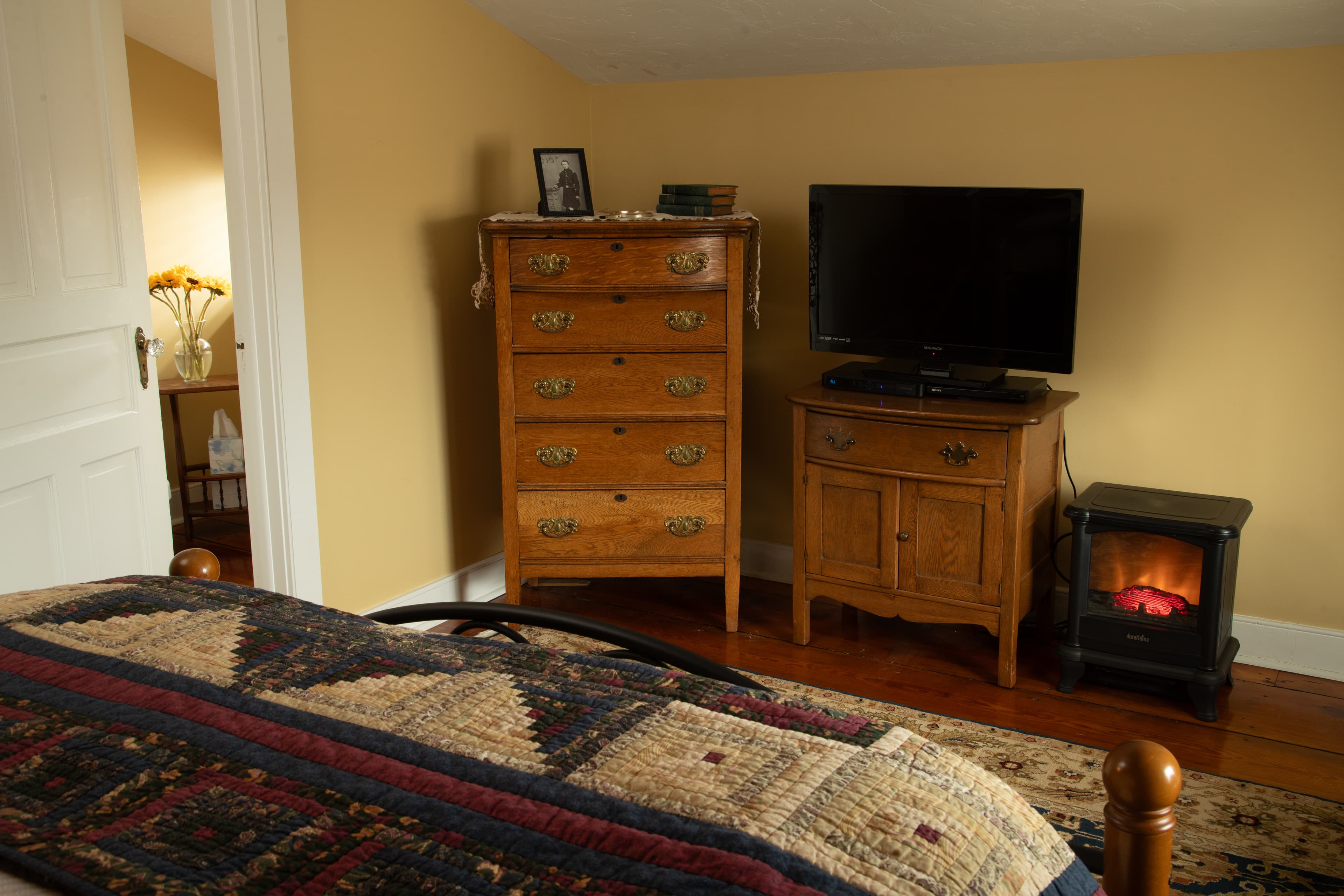 A cozy bedroom featuring a bed with a quilt, a wooden dresser, a TV, and a small electric heater against a warm-colored wall.