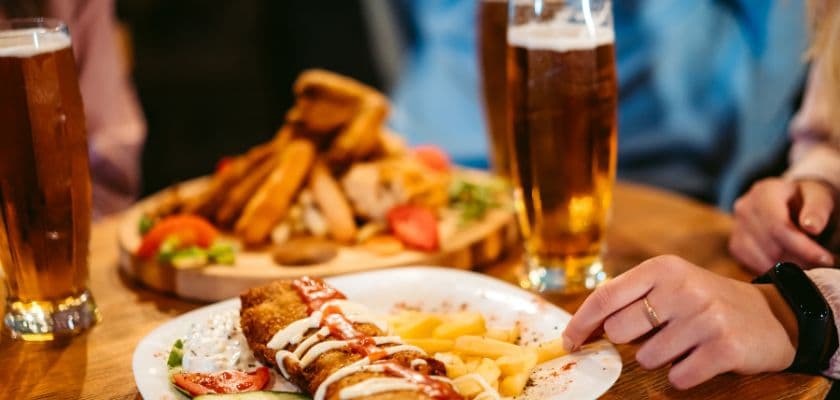 A hand reaches for a plate of fish and chips, accompanied by two glasses of beer and a shared platter of food.