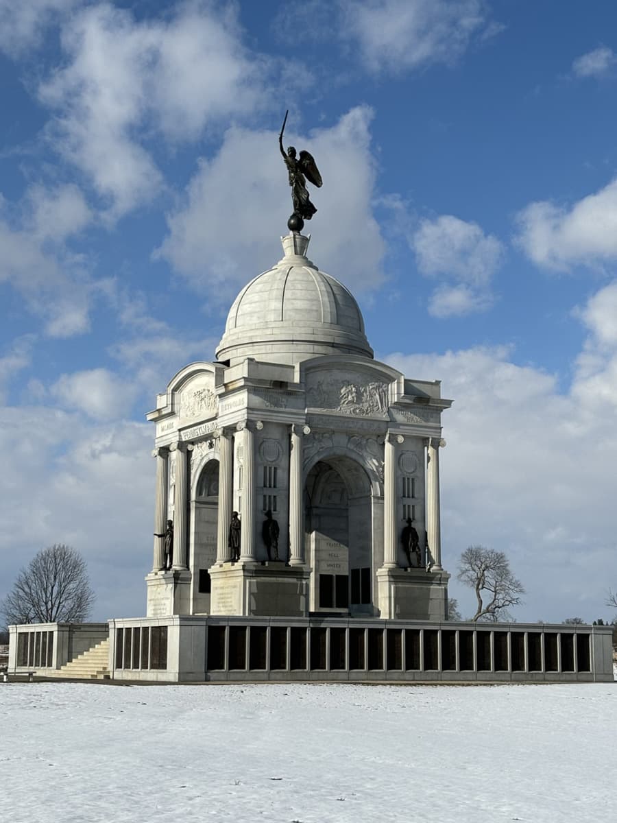 A monumental stone structure with a dome and a winged statue on top, set against a cloudy blue sky and surrounded by snow.