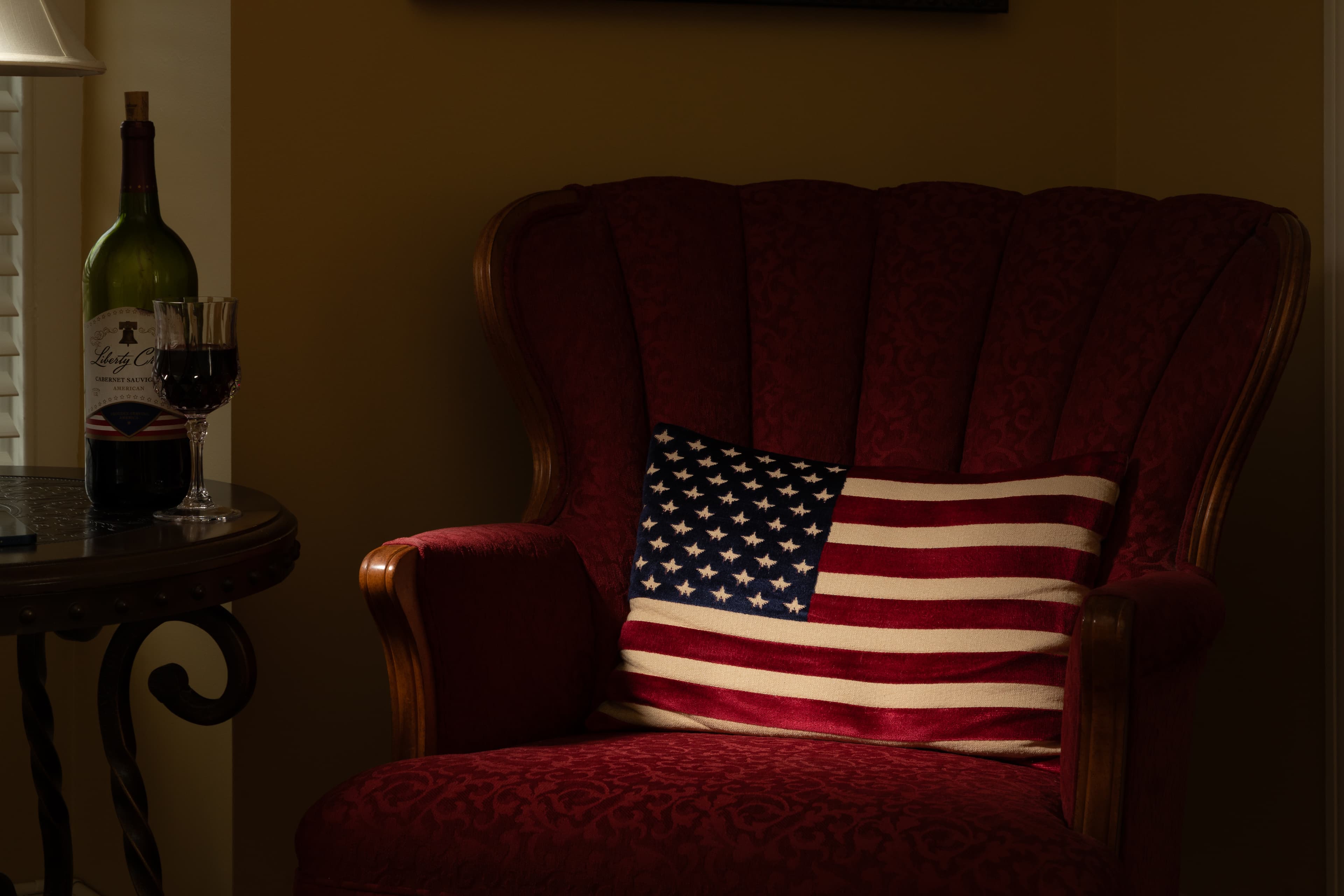 A red armchair with a striped American flag pillow next to a table holding a wine bottle and glass.