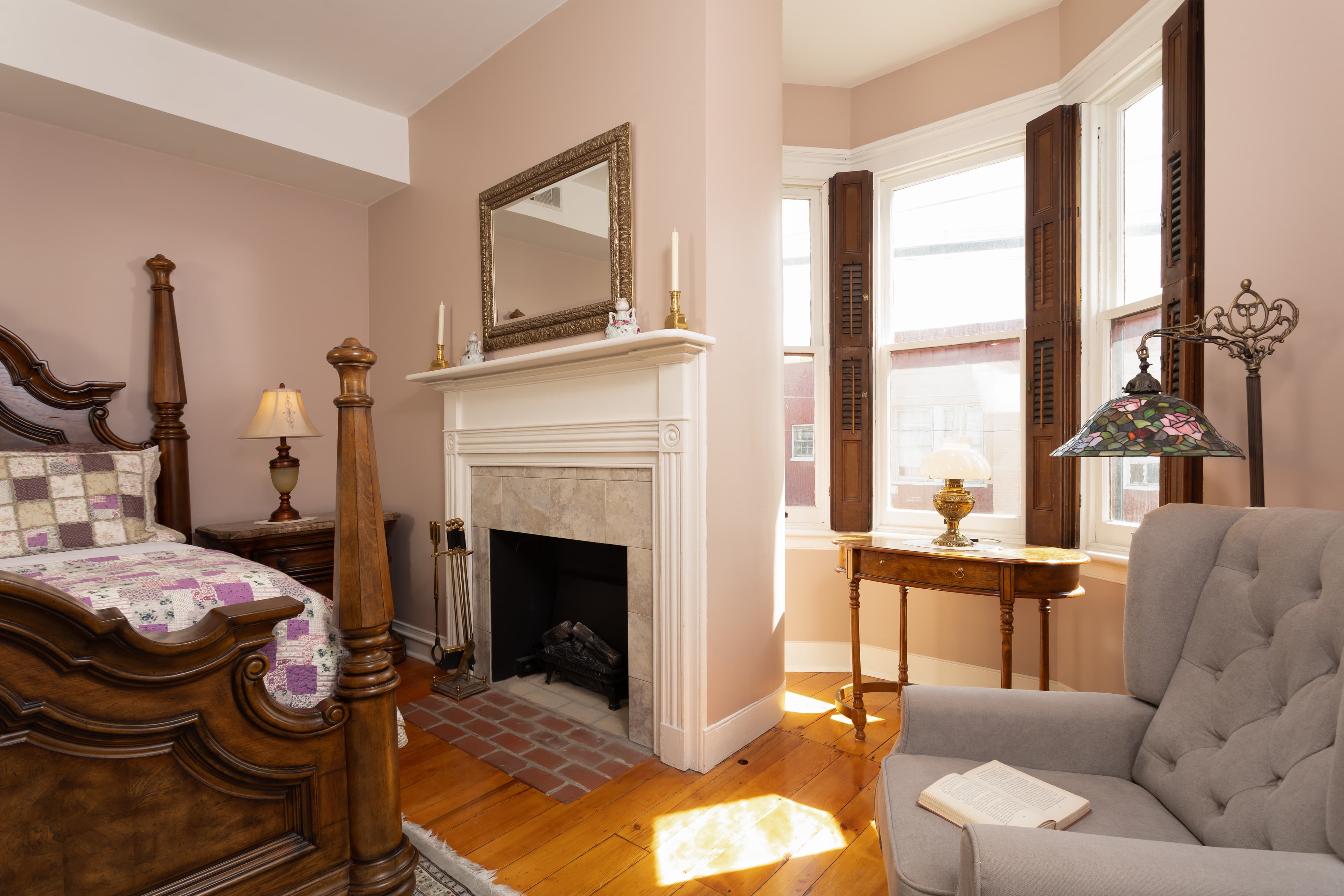 A cozy bedroom featuring a wooden bed, a fireplace, and a comfortable armchair beside a window.