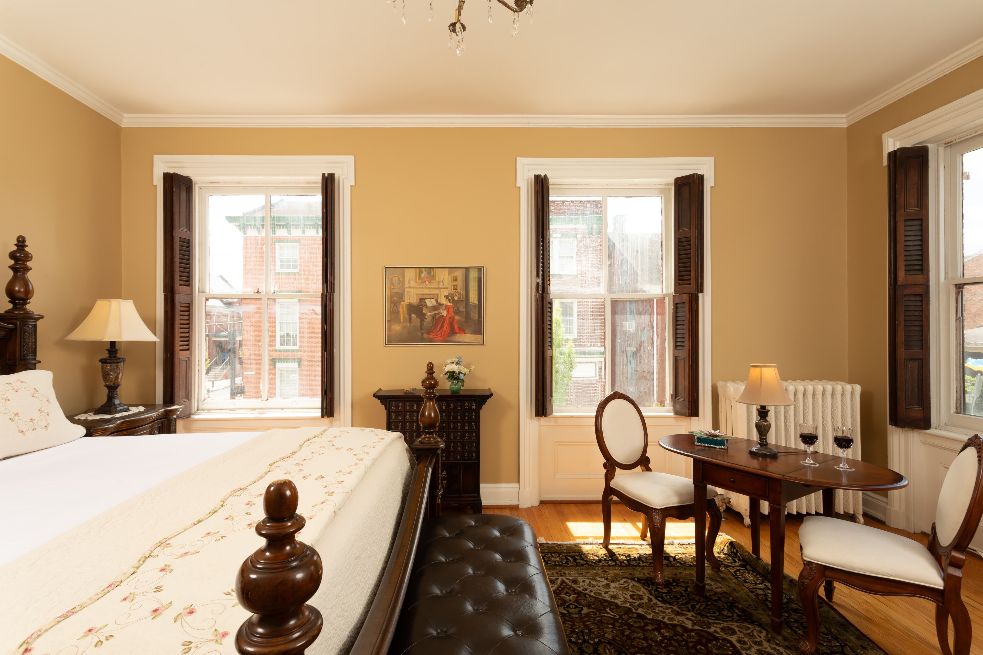 A cozy, elegantly furnished bedroom with a large bed, a small dining table, and natural light from two windows.