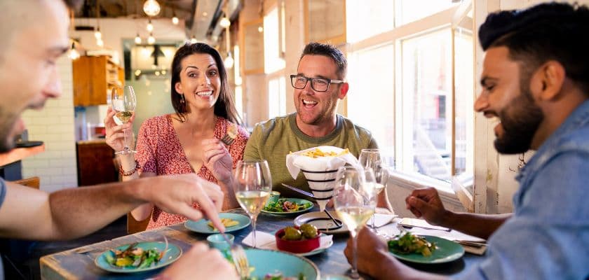 A group of four friends enjoys a lively meal together, laughing and toasting with drinks at a bright restaurant.